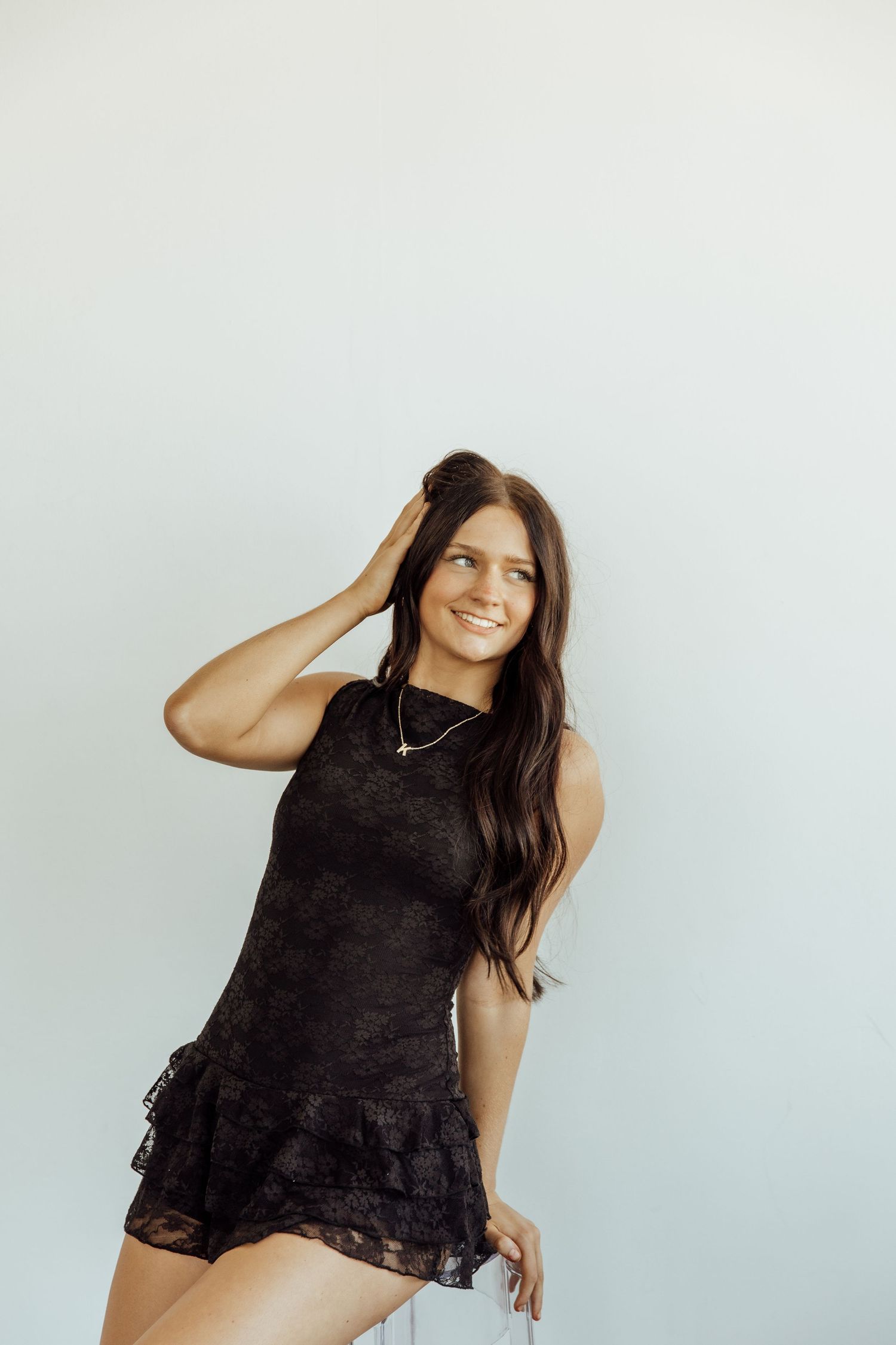 Smiling young woman in black lace mini dress posing against light gray background with hand in hair.