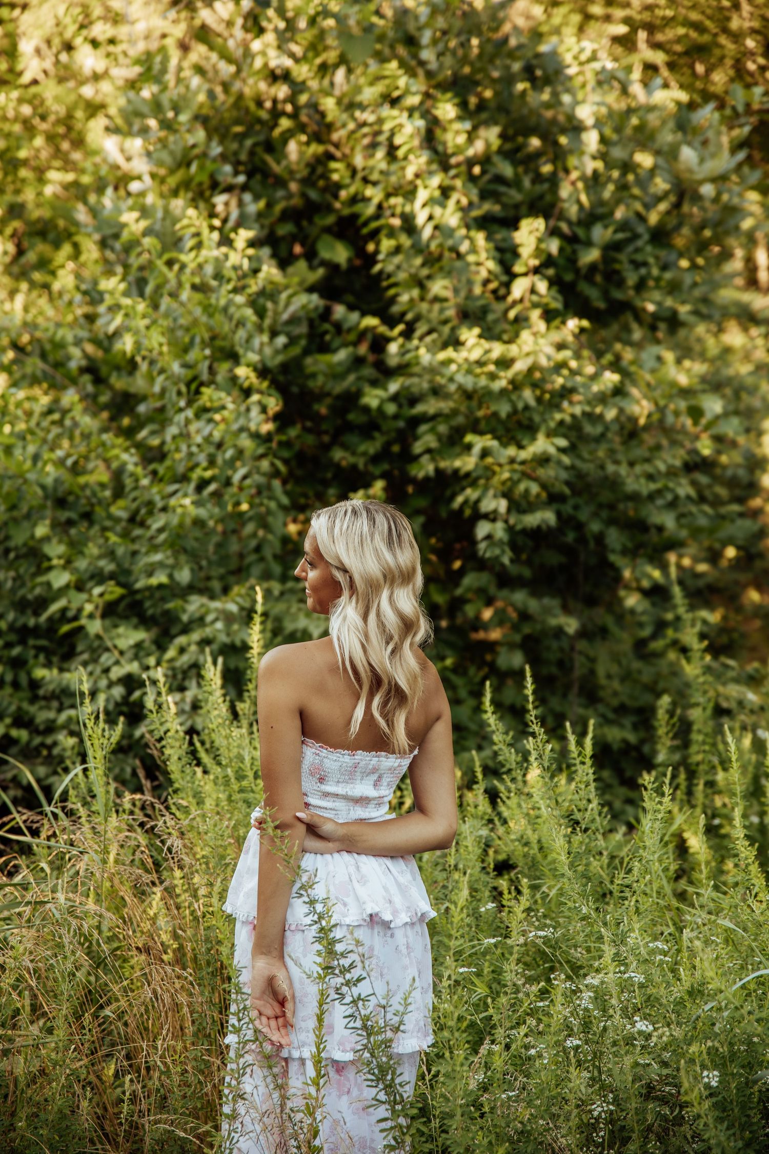 Blonde woman in white floral strapless dress standing with back turned in lush green garden.
