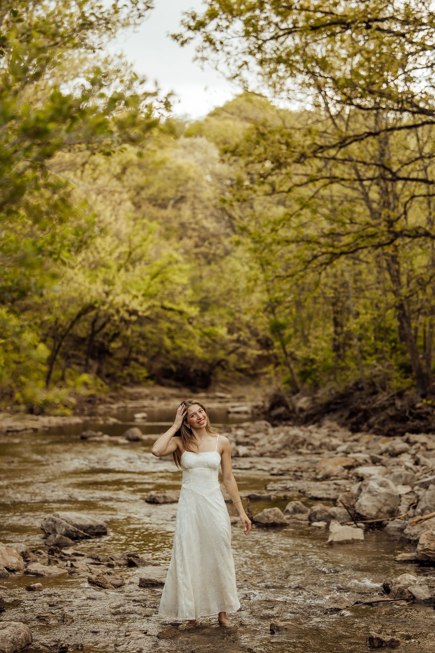 Young woman in elegant white dress standing in rocky creek surrounded by lush green forest.