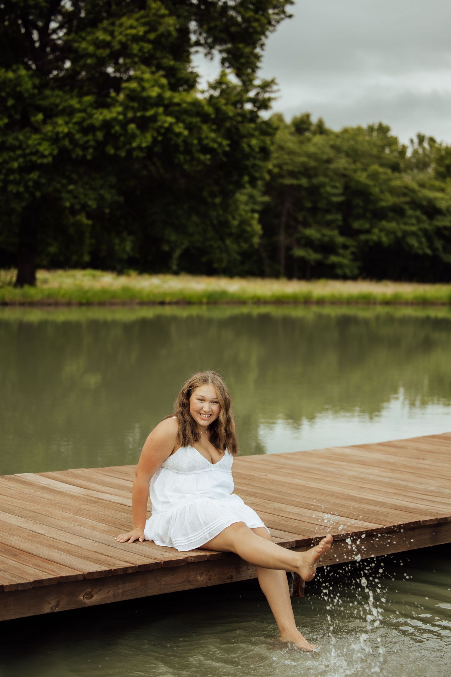 Young woman in white dress sitting on wooden dock, splashing feet in calm lake surrounded by lush green trees.