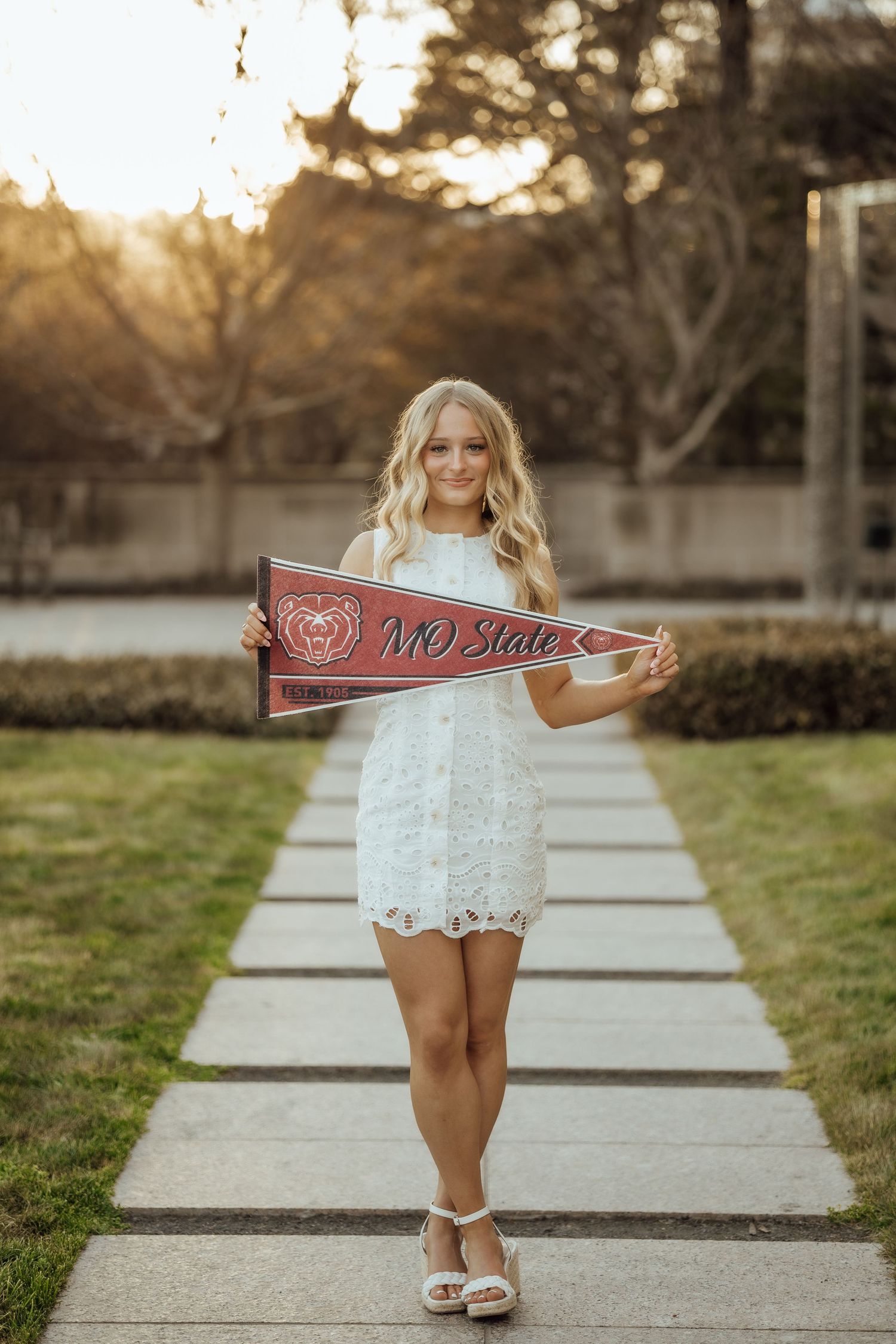 Smiling blonde in white lace dress holds MO State pennant on sunlit garden path.
