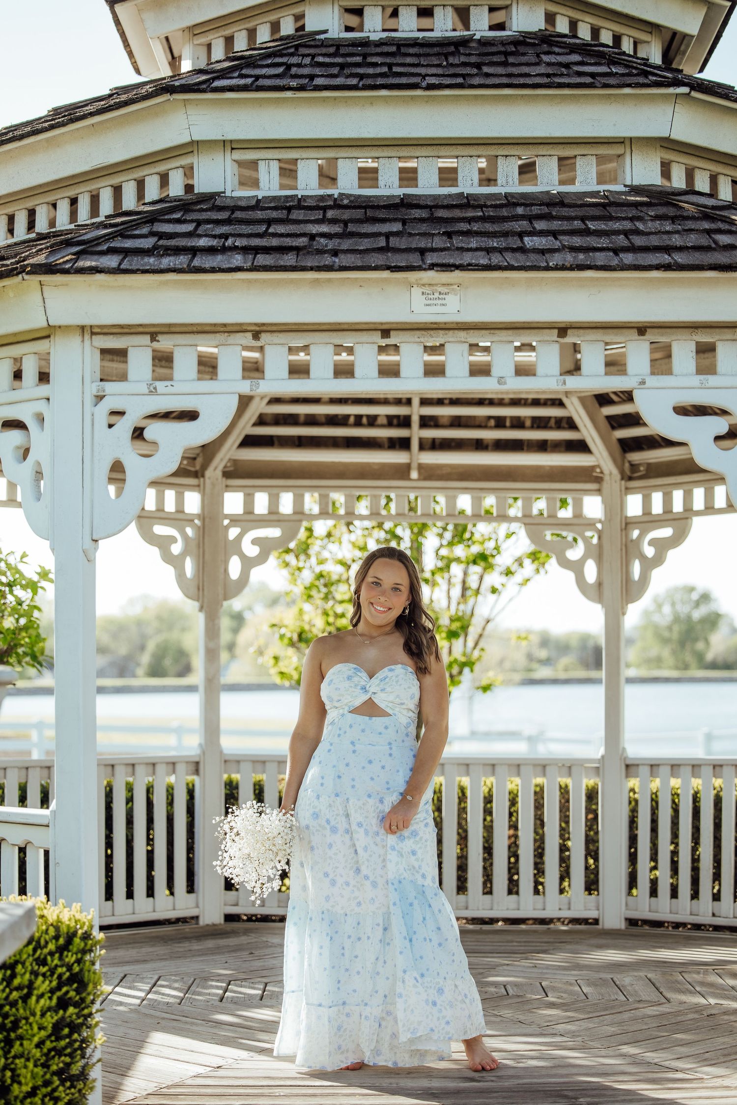 Young woman in blue floral dress holding white bouquet standing inside white gazebo near water.