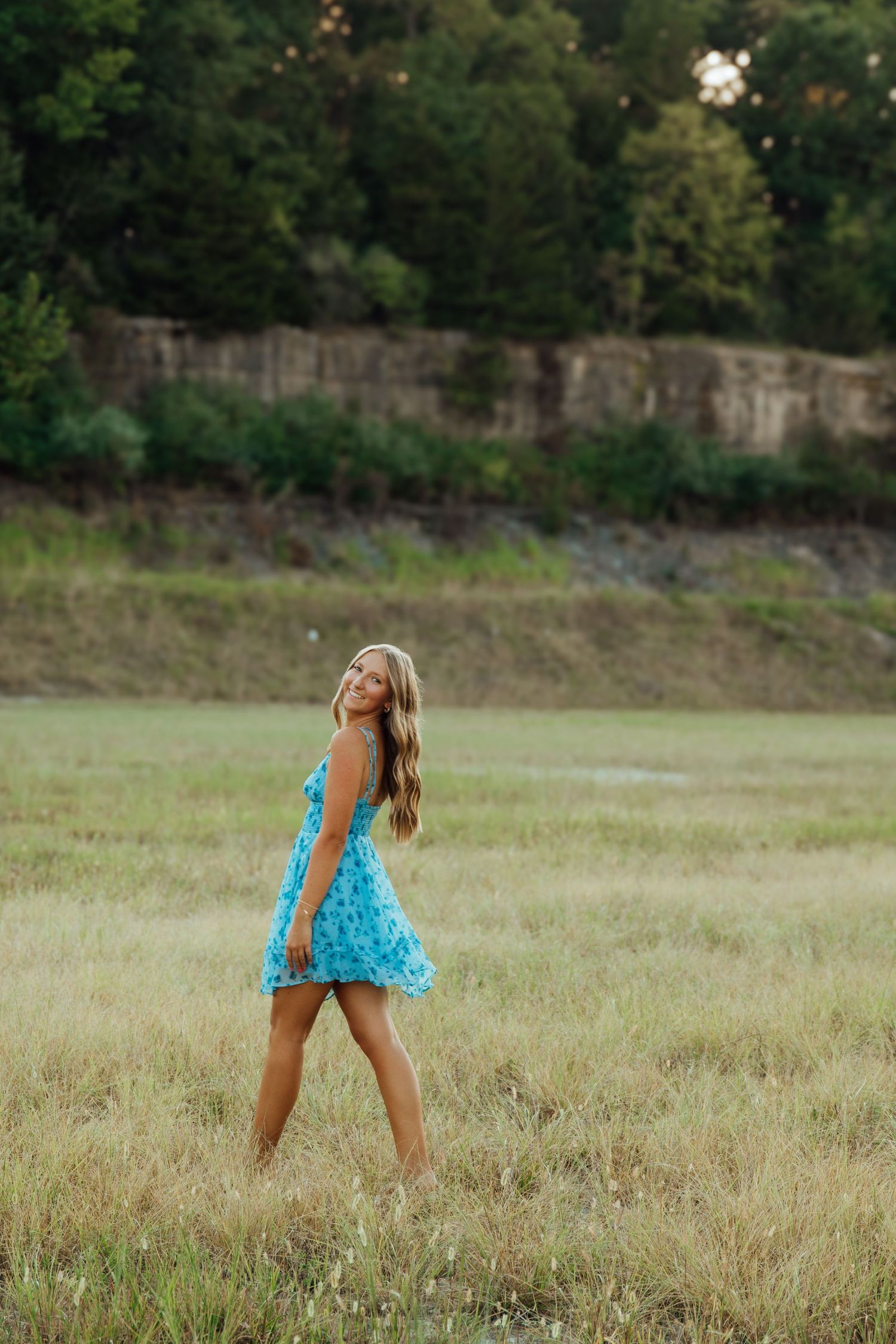 Young woman in blue floral sundress walking through a grassy field at golden hour.