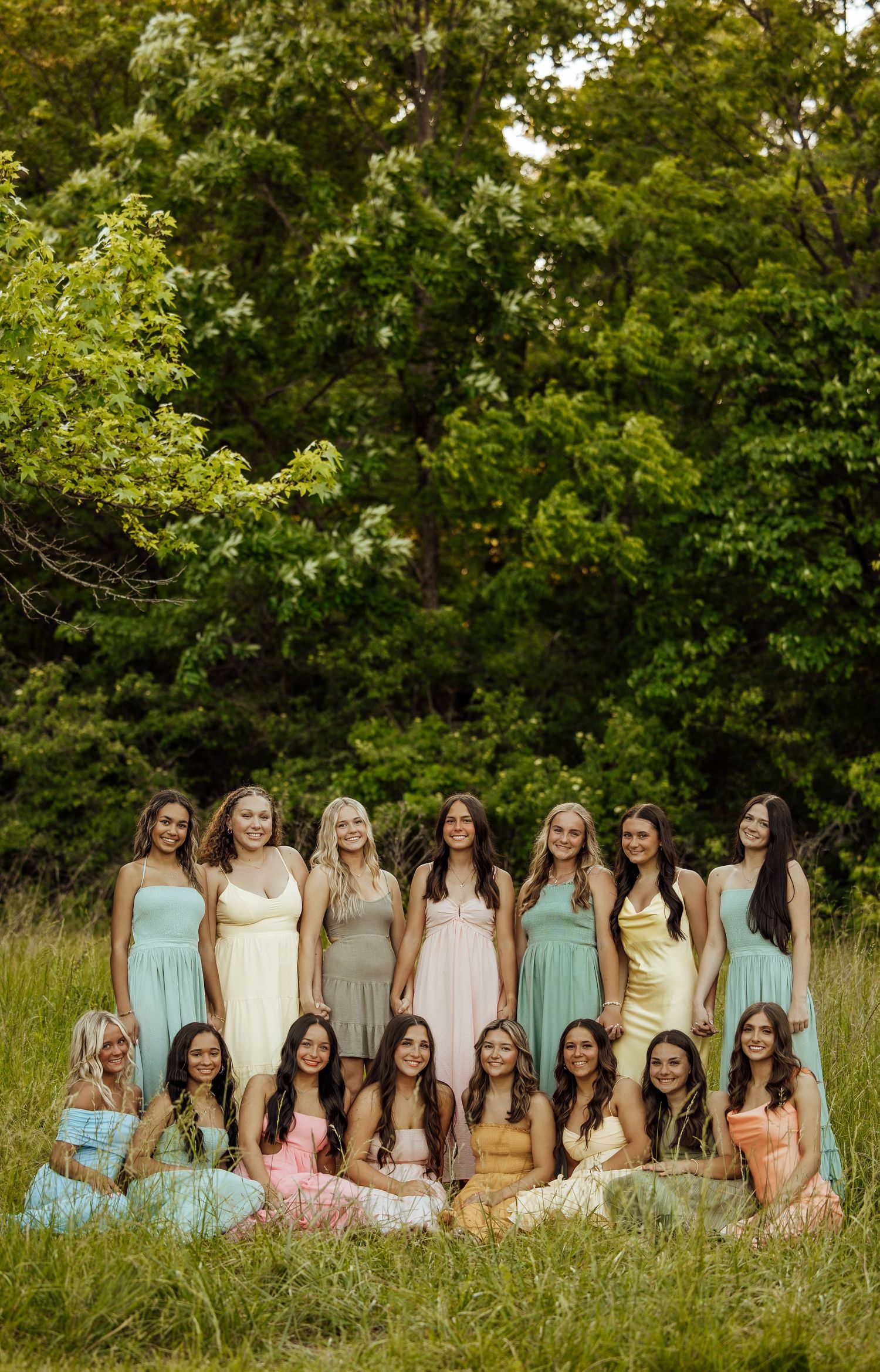 Large group of young women in colorful pastel dresses posing outdoors in green grassy field.