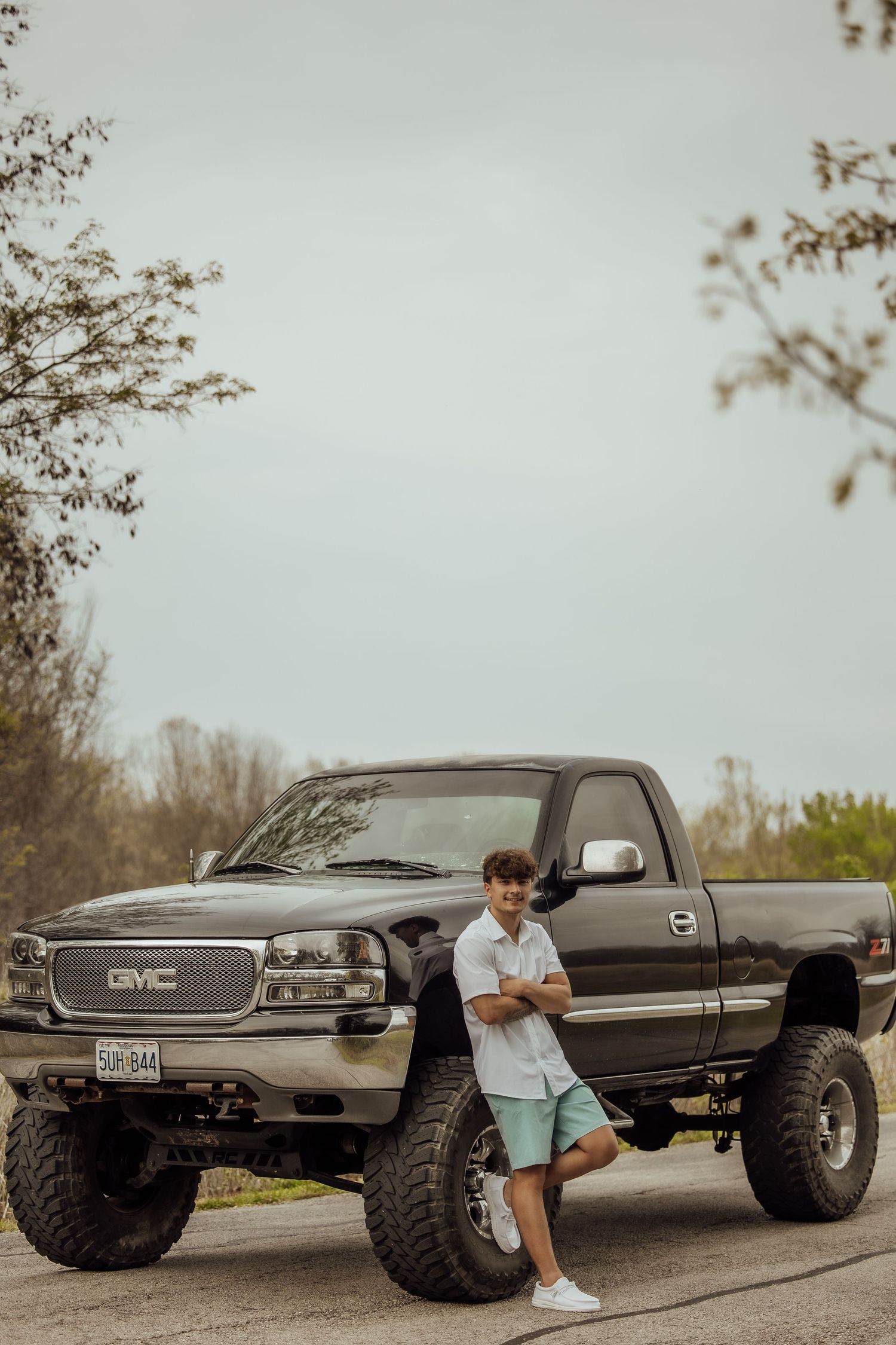 Teen boy in white shirt poses leaning against lifted GMC pickup truck outdoors.