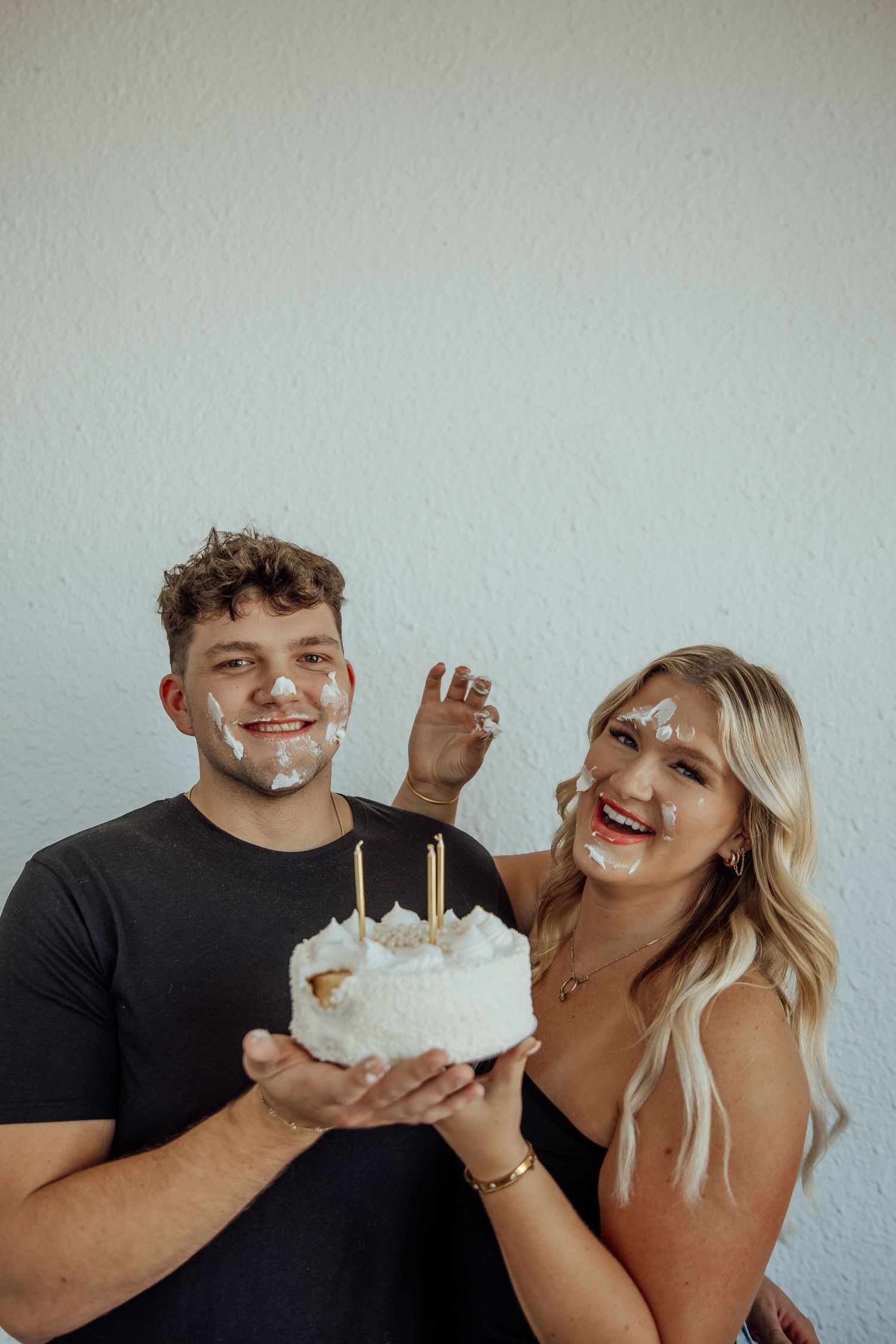 A smiling couple holds a white birthday cake with candles, both with frosting on their faces, celebrating together.