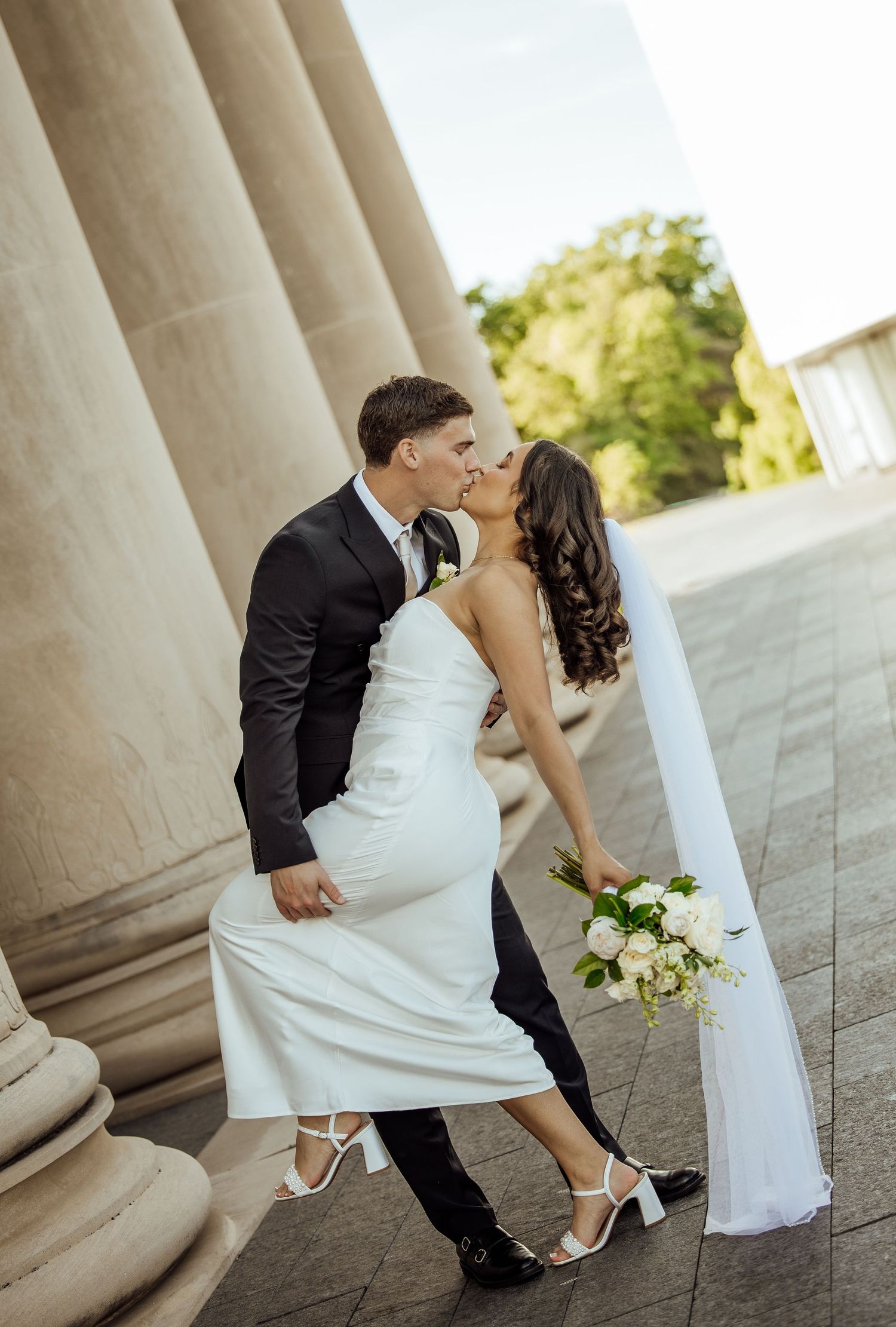Bride and groom sharing a romantic kiss beside tall stone columns on their wedding day.