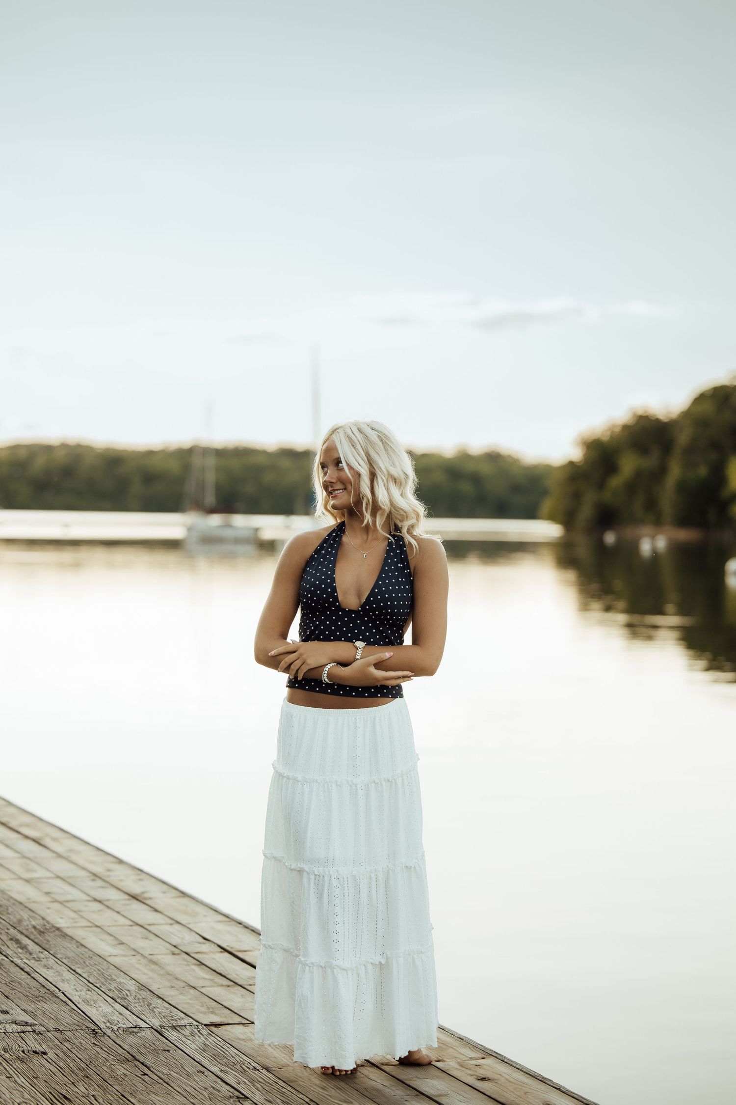 Blonde woman in polka dot halter top and white maxi skirt standing on wooden dock by calm lake at sunset.