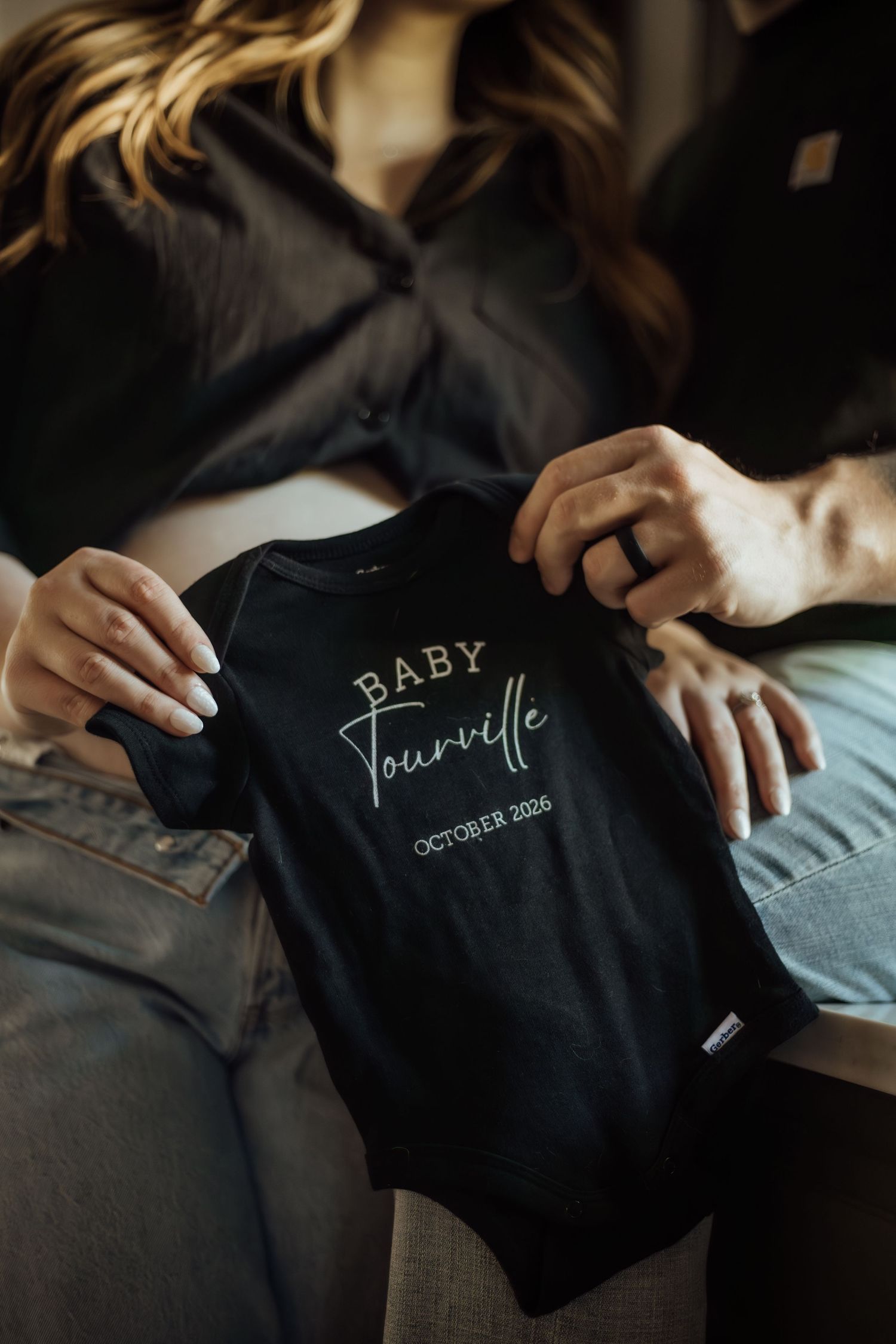 A couple holds a black baby onesie reading Baby Tourville October 2026 in a cozy indoor pregnancy announcement photo.