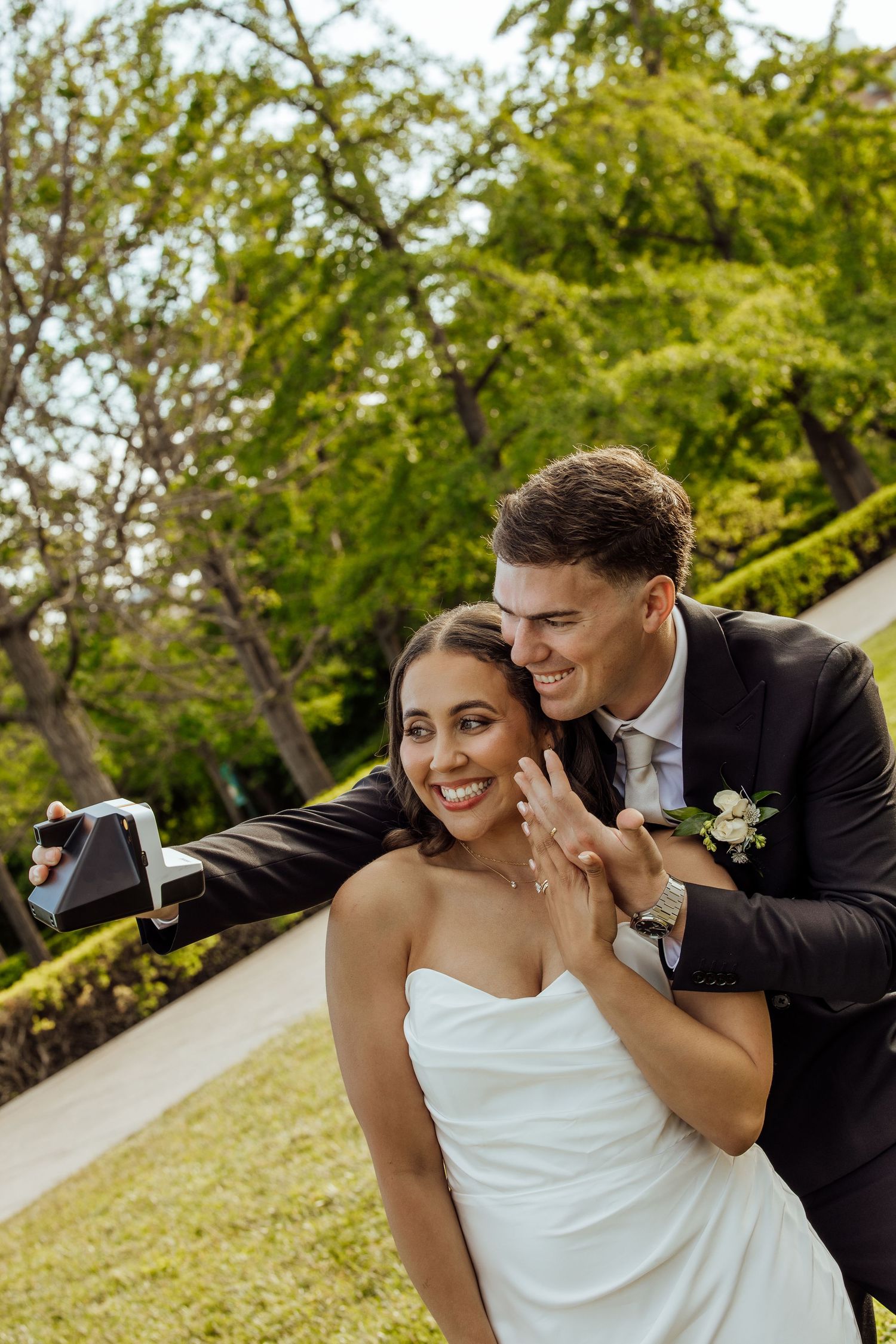 Happy bride and groom taking a Polaroid selfie outdoors, bride showing off her wedding ring.
