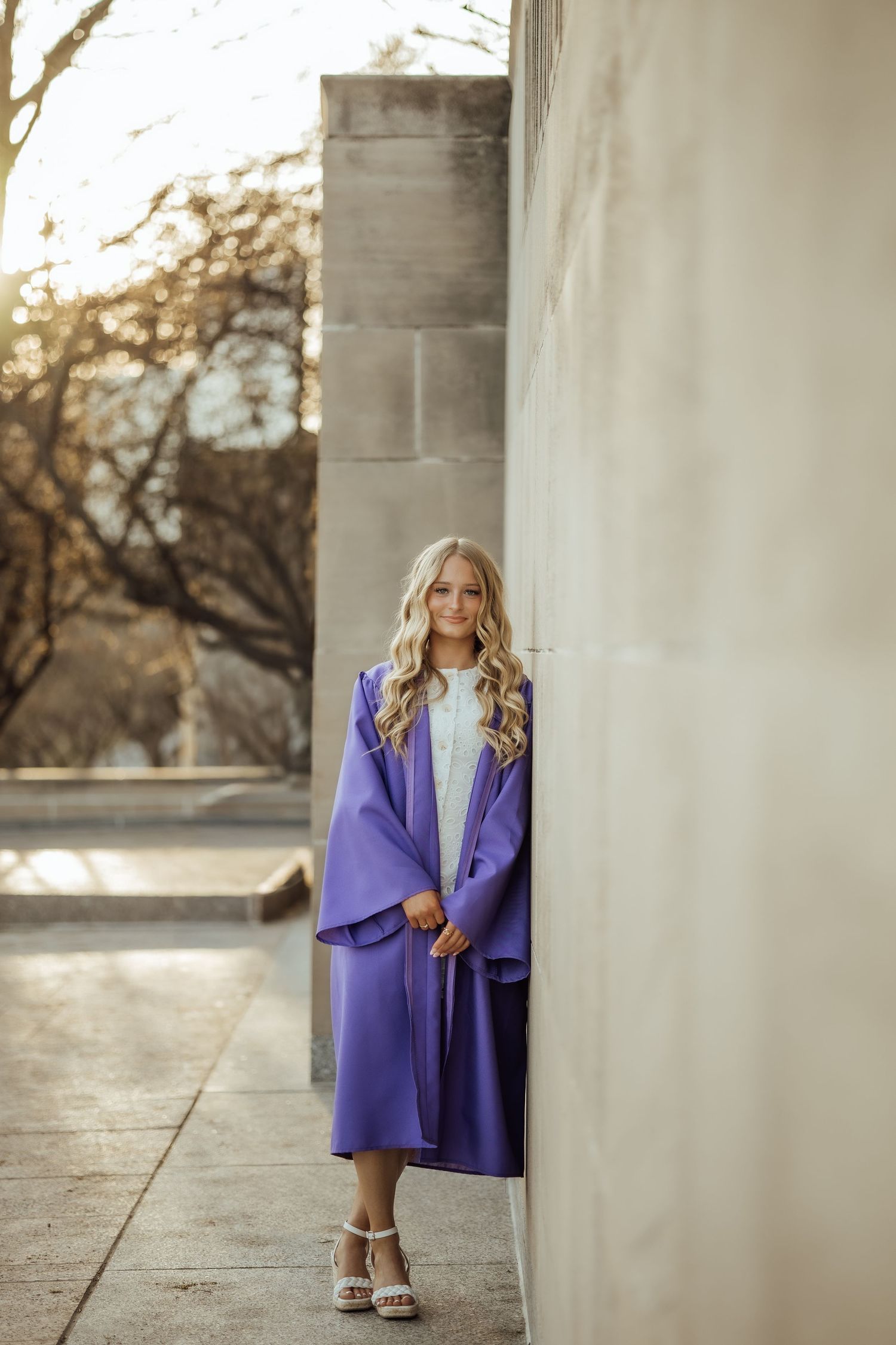 Blonde graduate in purple cap and gown leans against stone wall in warm sunlight.