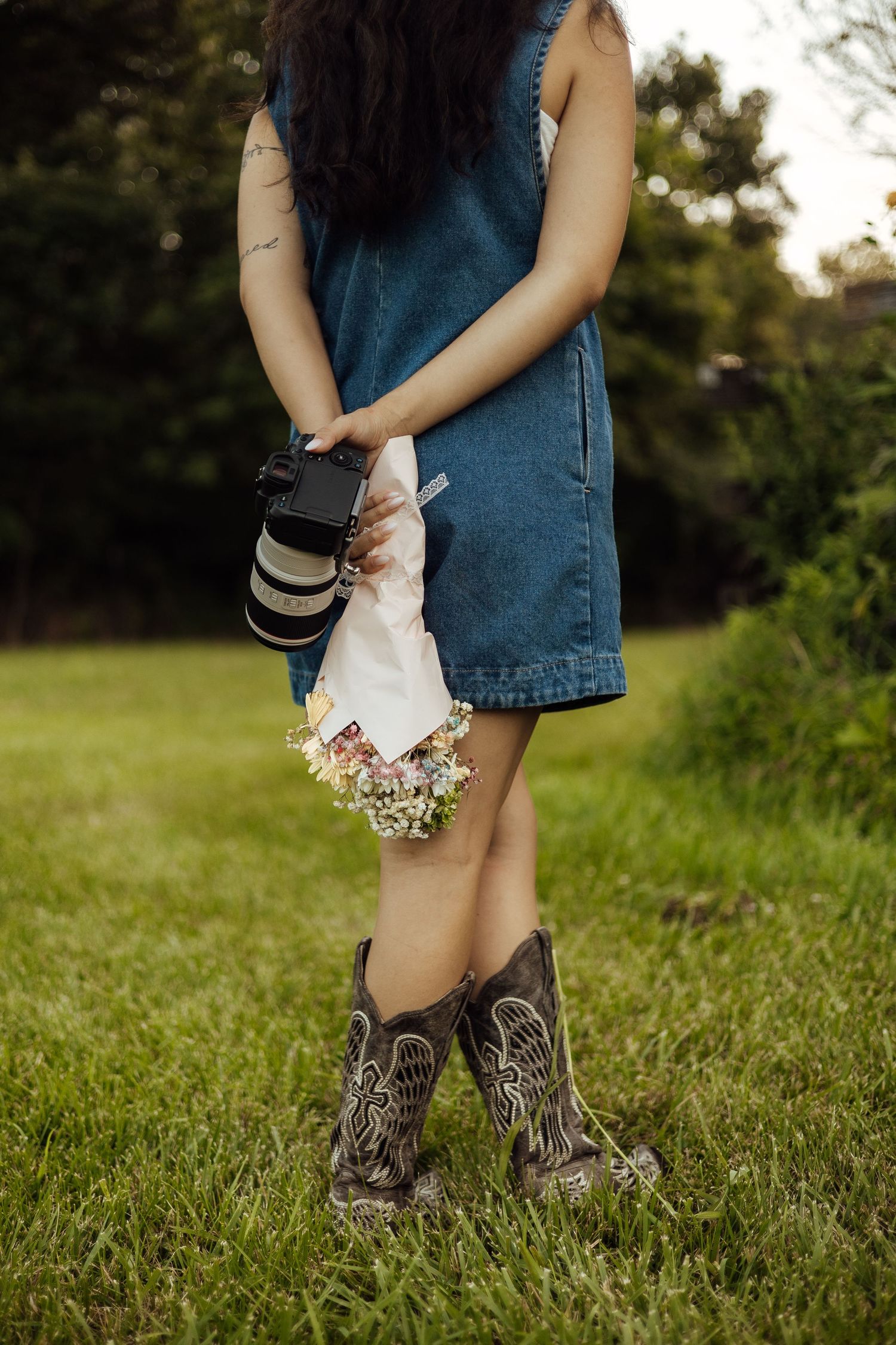 Woman in denim dress and cowboy boots holding a camera and wildflowers while standing on green grass outdoors.