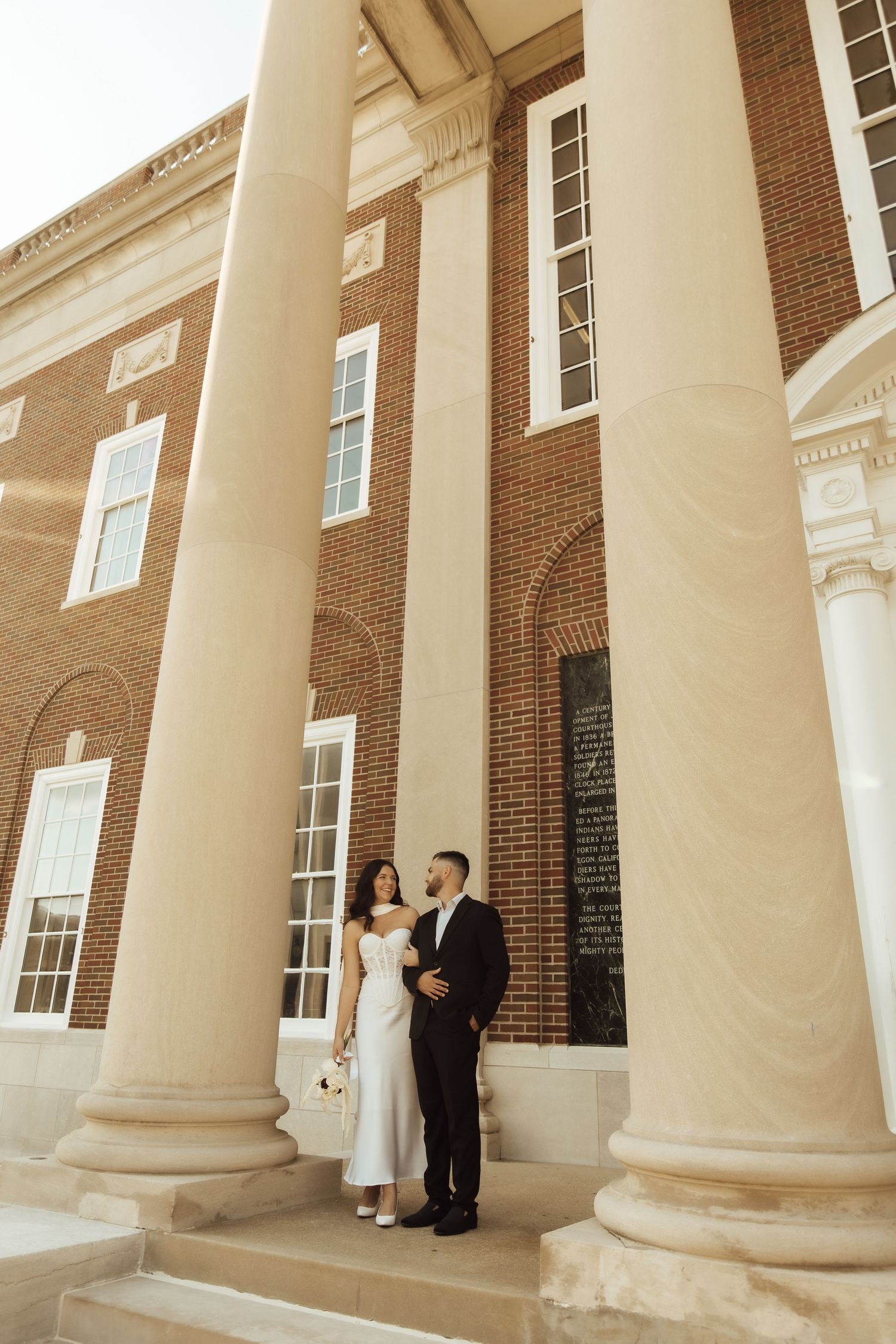Bride and groom posing between white columns of a brick building on their wedding day.