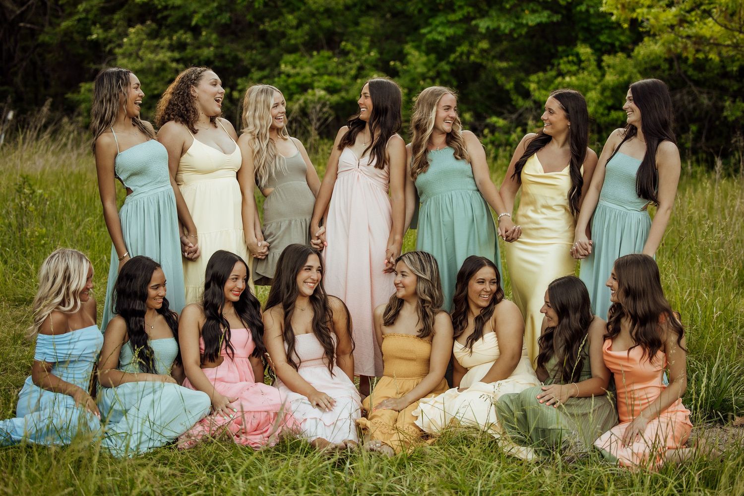 Group of young women in elegant pastel gowns laughing together in a sunlit grassy meadow.