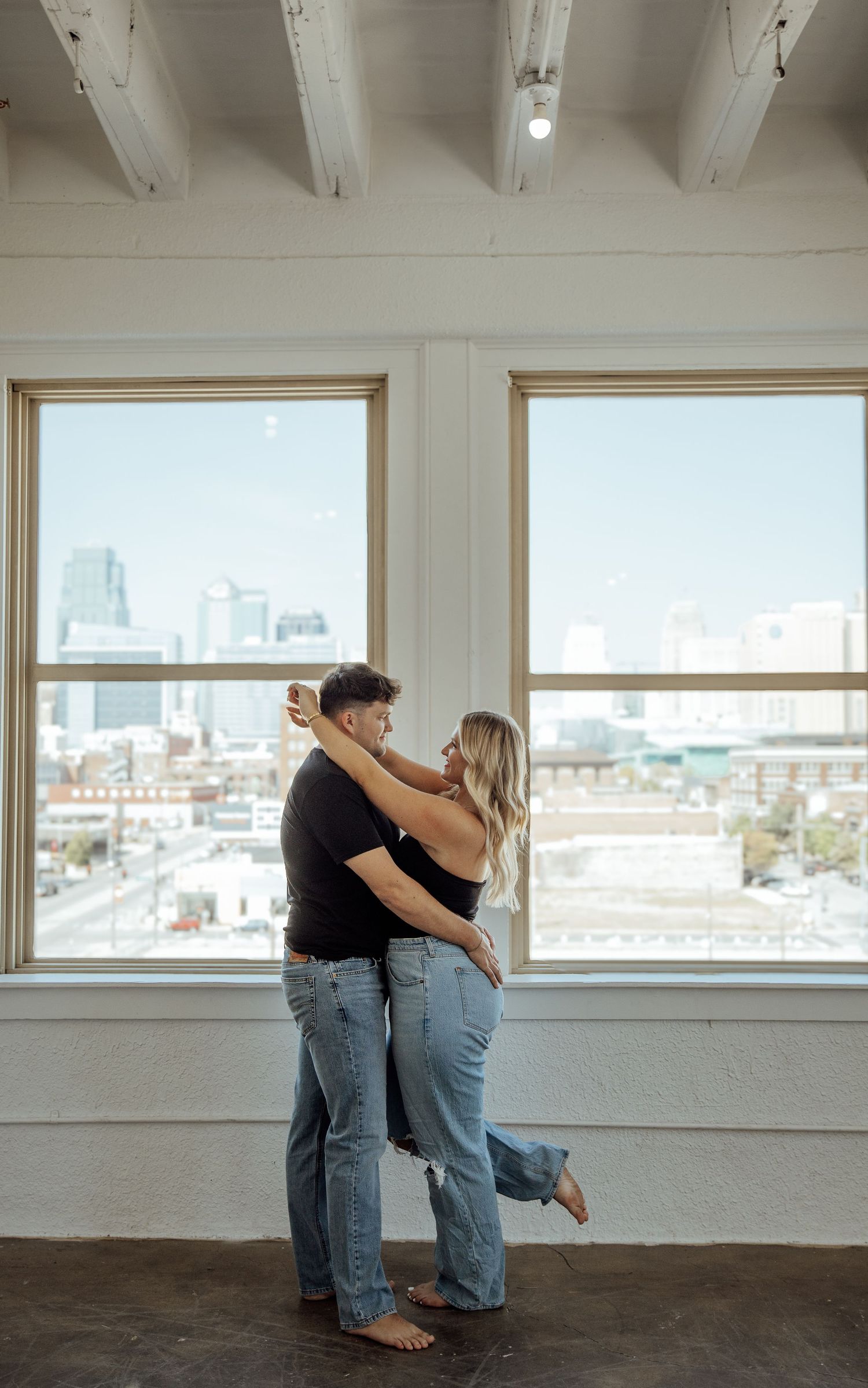 Couple dancing and embracing near large windows with city skyline view, both wearing black tops and jeans.