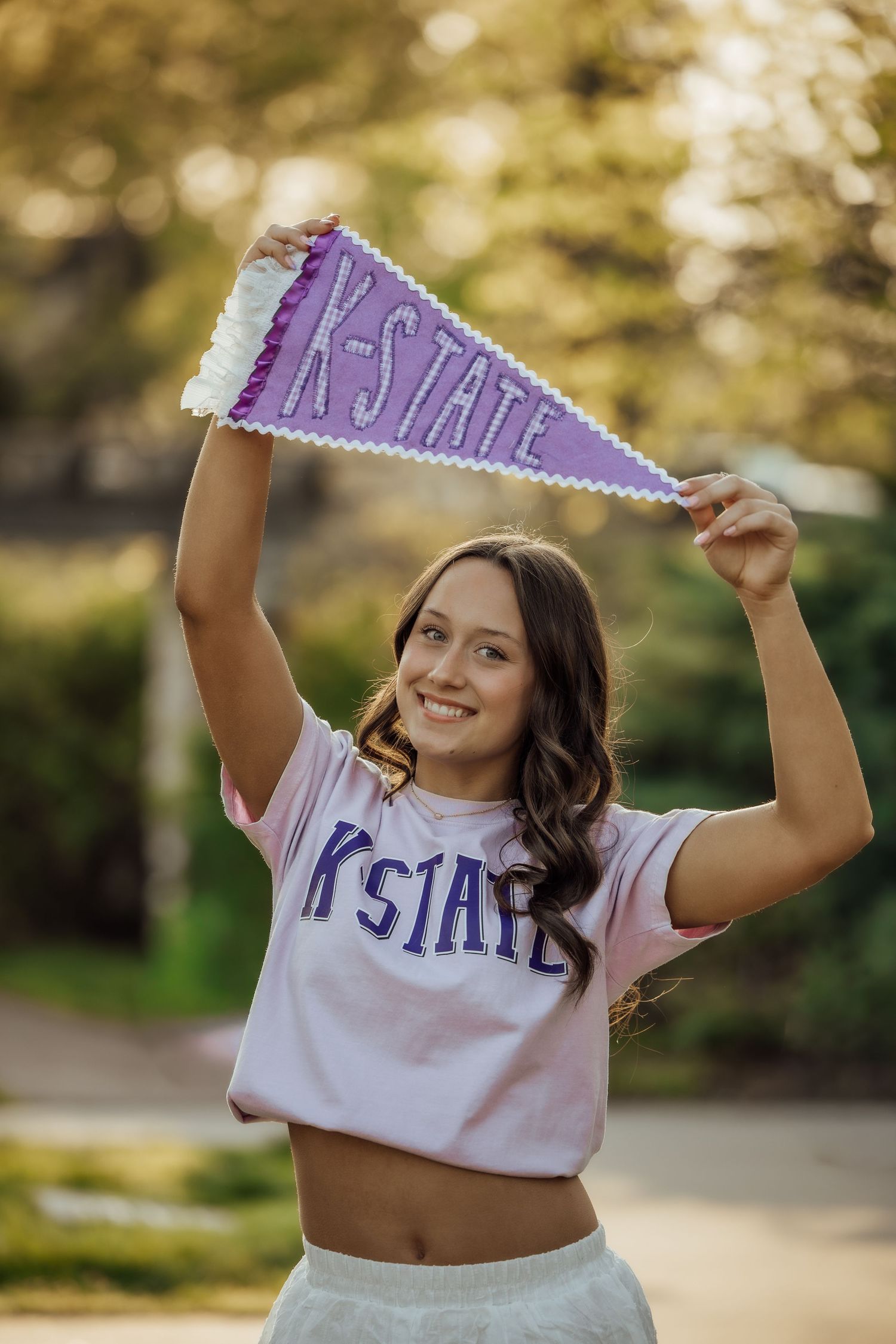 Brunette woman in K-State shirt raises purple pennant overhead in outdoor setting.