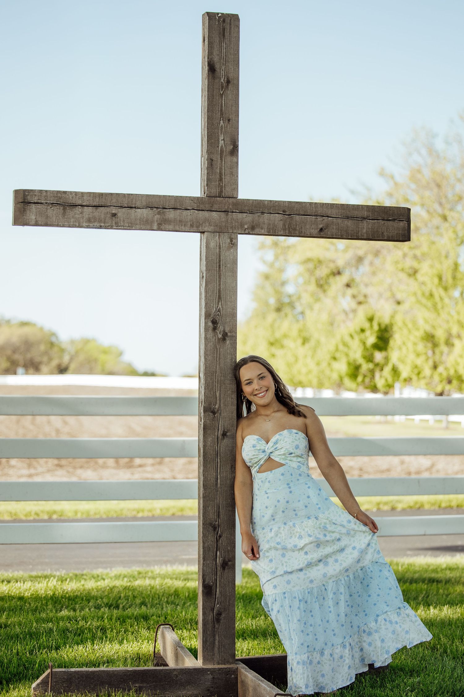 Smiling young woman in blue floral dress leaning against large wooden cross on green lawn.