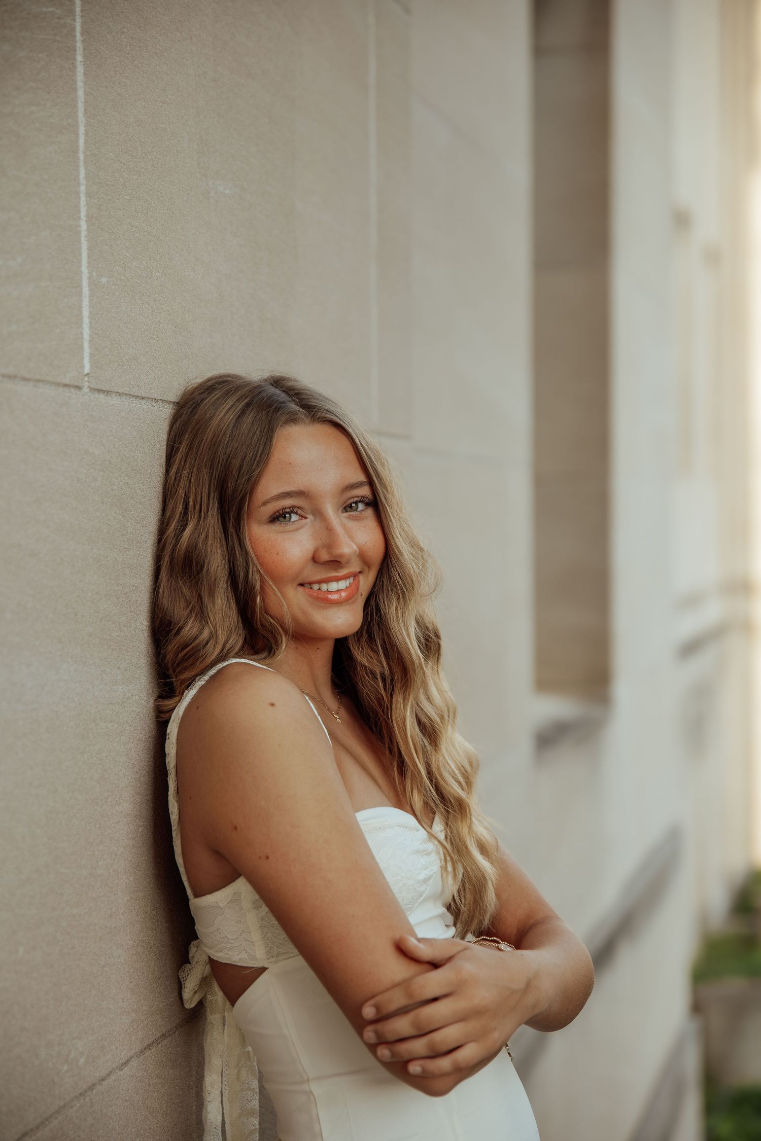 Young blonde woman in white dress leaning against stone wall smiling softly in warm golden light.