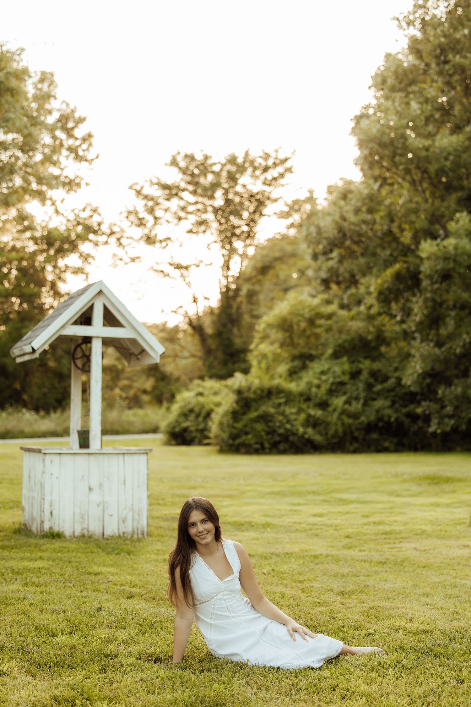 Young woman in white dress sitting on green grass near a vintage wooden wishing well in a sunny park.