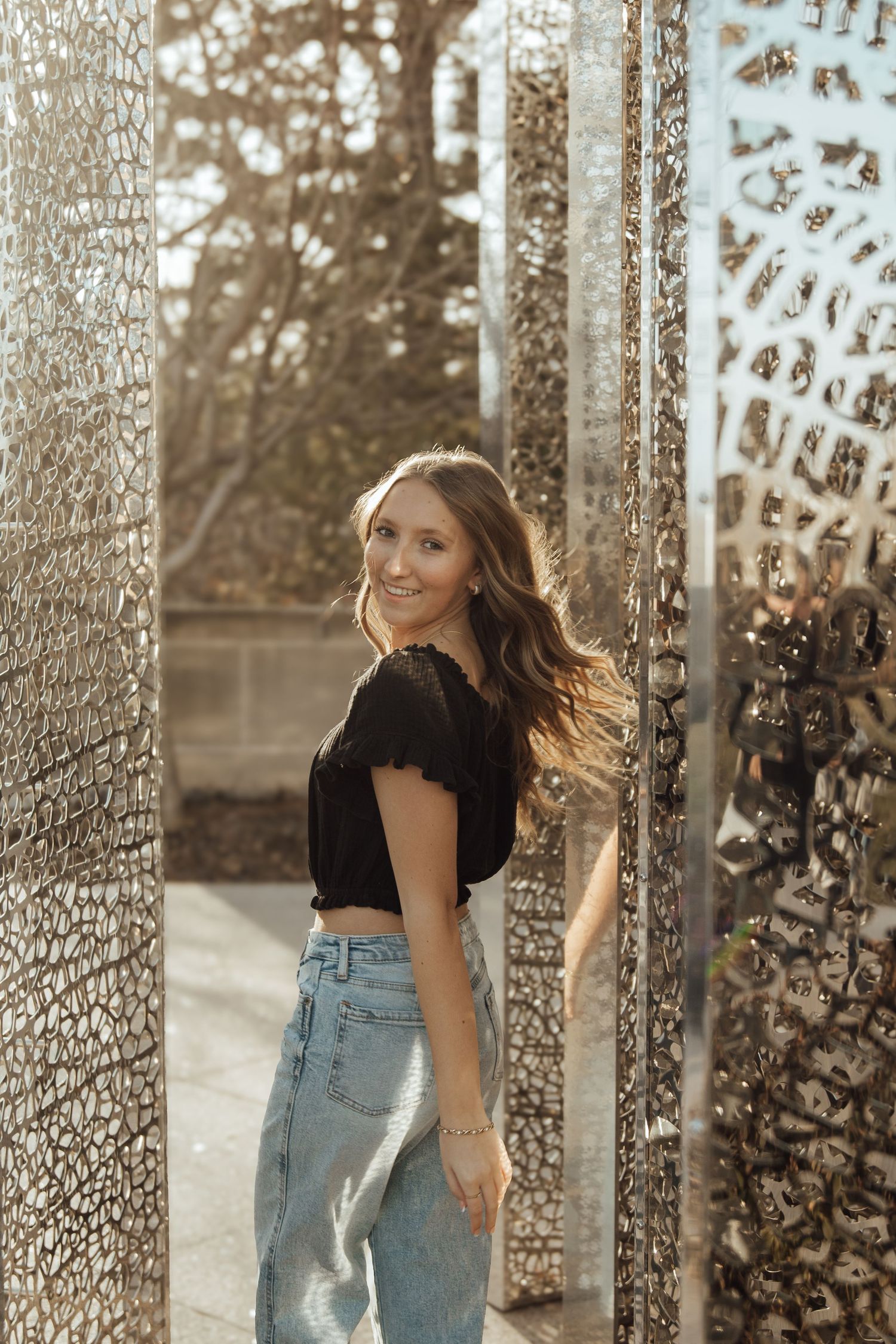 Smiling young woman in black crop top and jeans posing between decorative metal lattice panels.