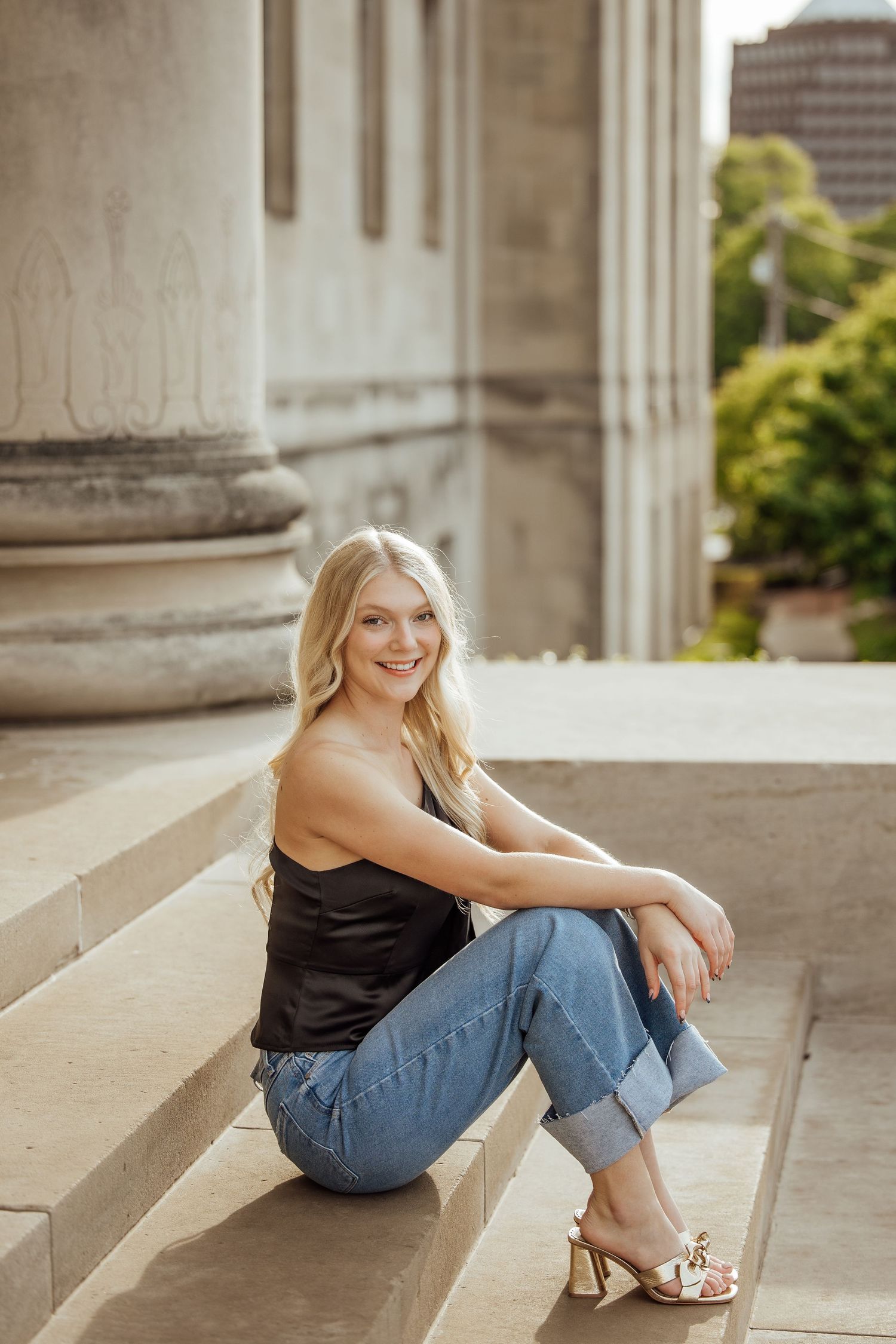 Blonde woman in black camisole and jeans sits smiling on stone steps near columns.