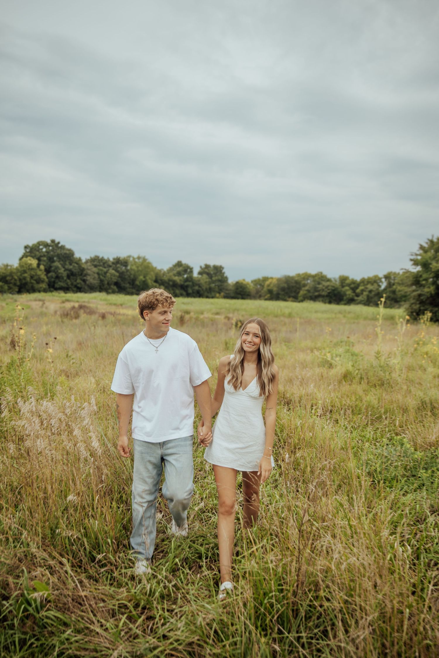 A young couple holds hands while walking through a lush green meadow under a cloudy sky.