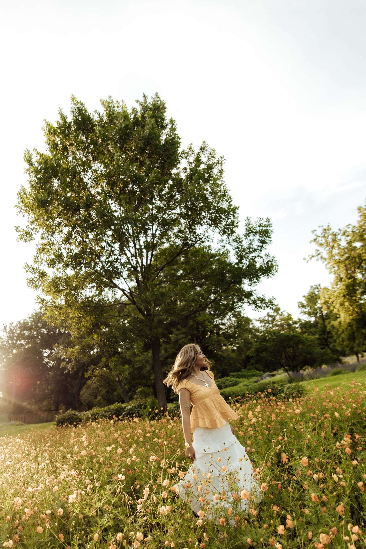 Woman in white floral dress walking through a sunlit meadow with wildflowers and lush green trees in the background.
