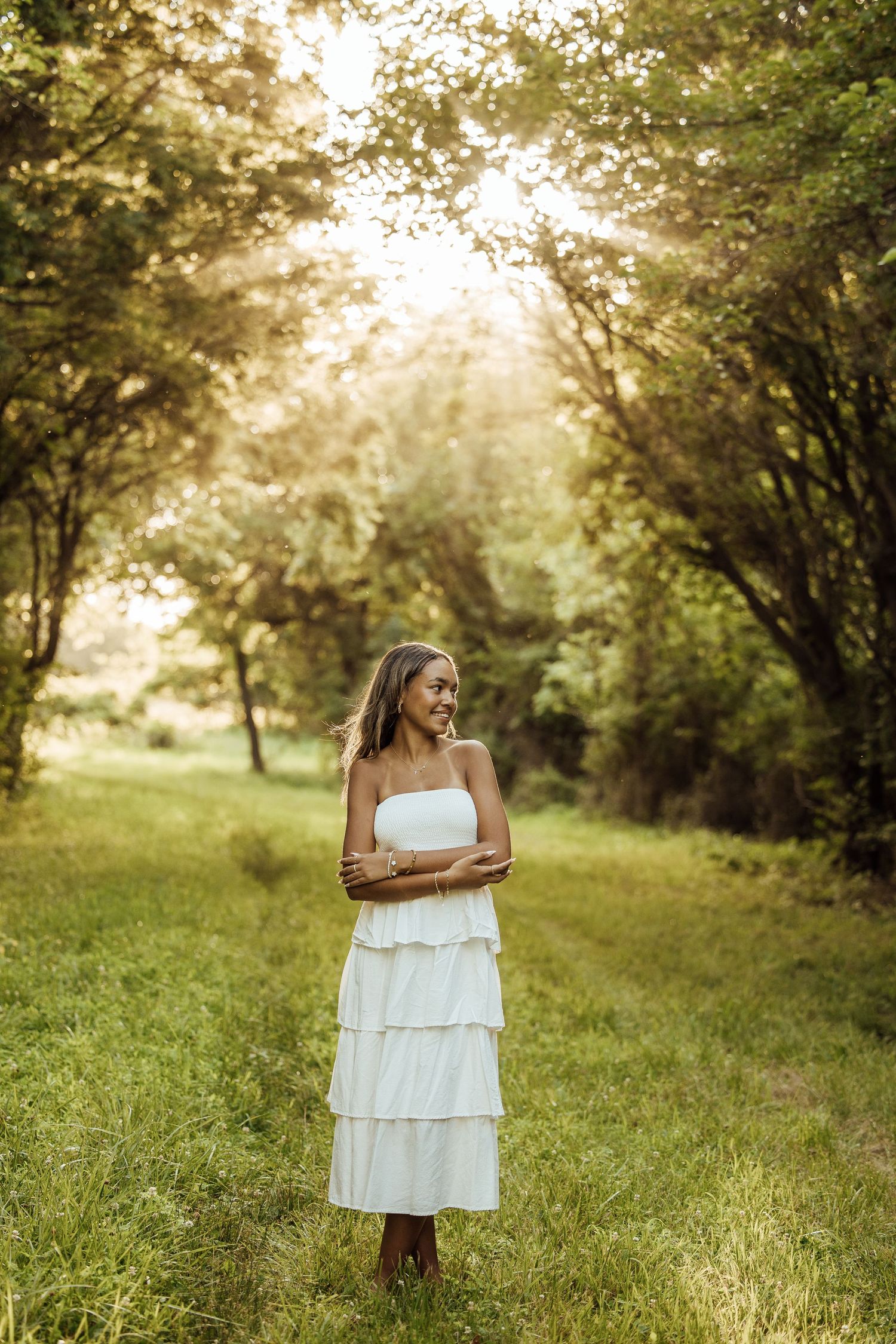 Young woman in white tiered strapless dress smiling in sunlit green forest path, golden hour outdoor portrait.