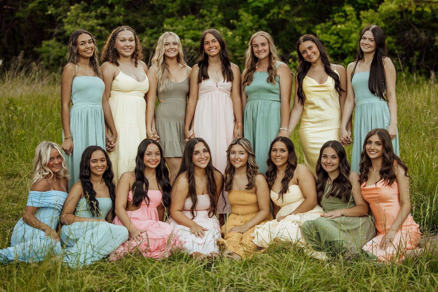 Bridesmaids in pastel dresses smiling together in two rows on green grass.