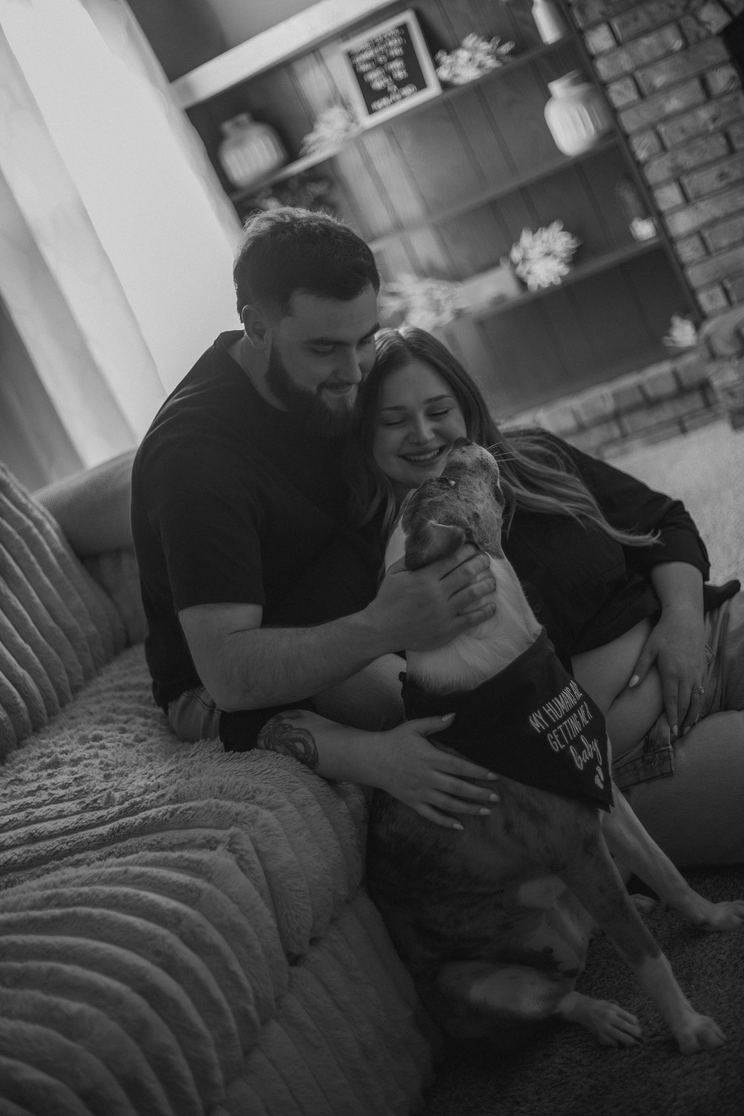A happy couple cuddles with their dog on a couch in a cozy living room in a black and white portrait.