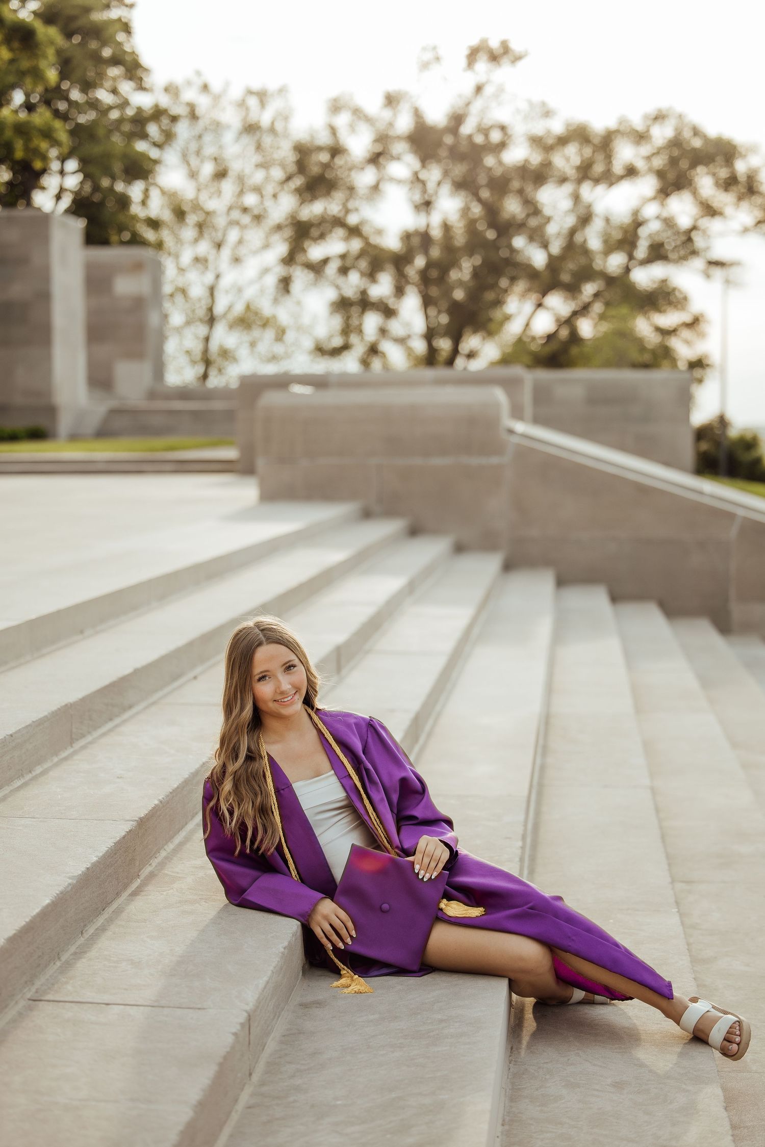 Young woman in purple graduation cap and gown sitting on stone steps outdoors at golden hour.