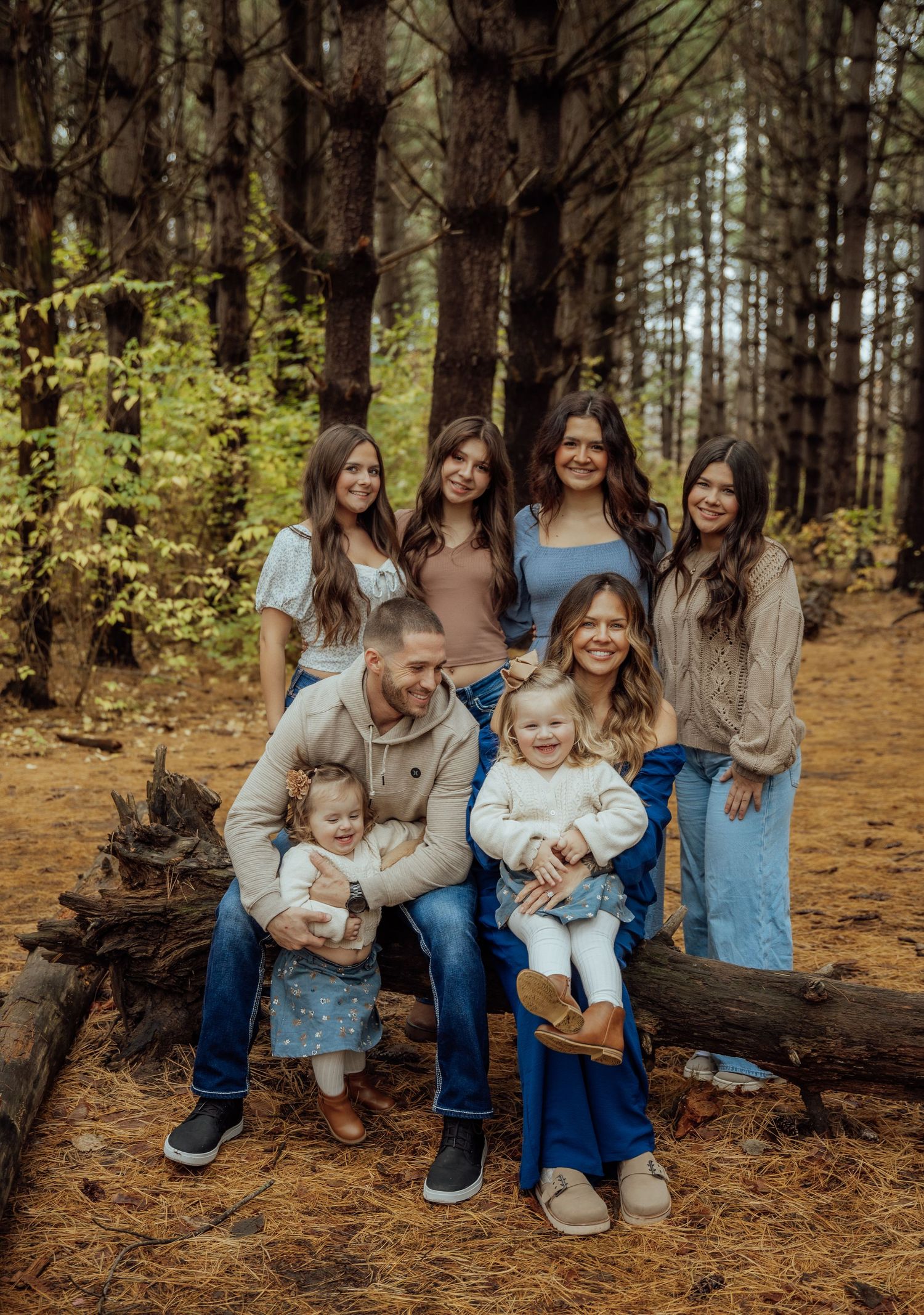 Blended family of seven posing on a fallen log in a pine forest during an outdoor family portrait session.