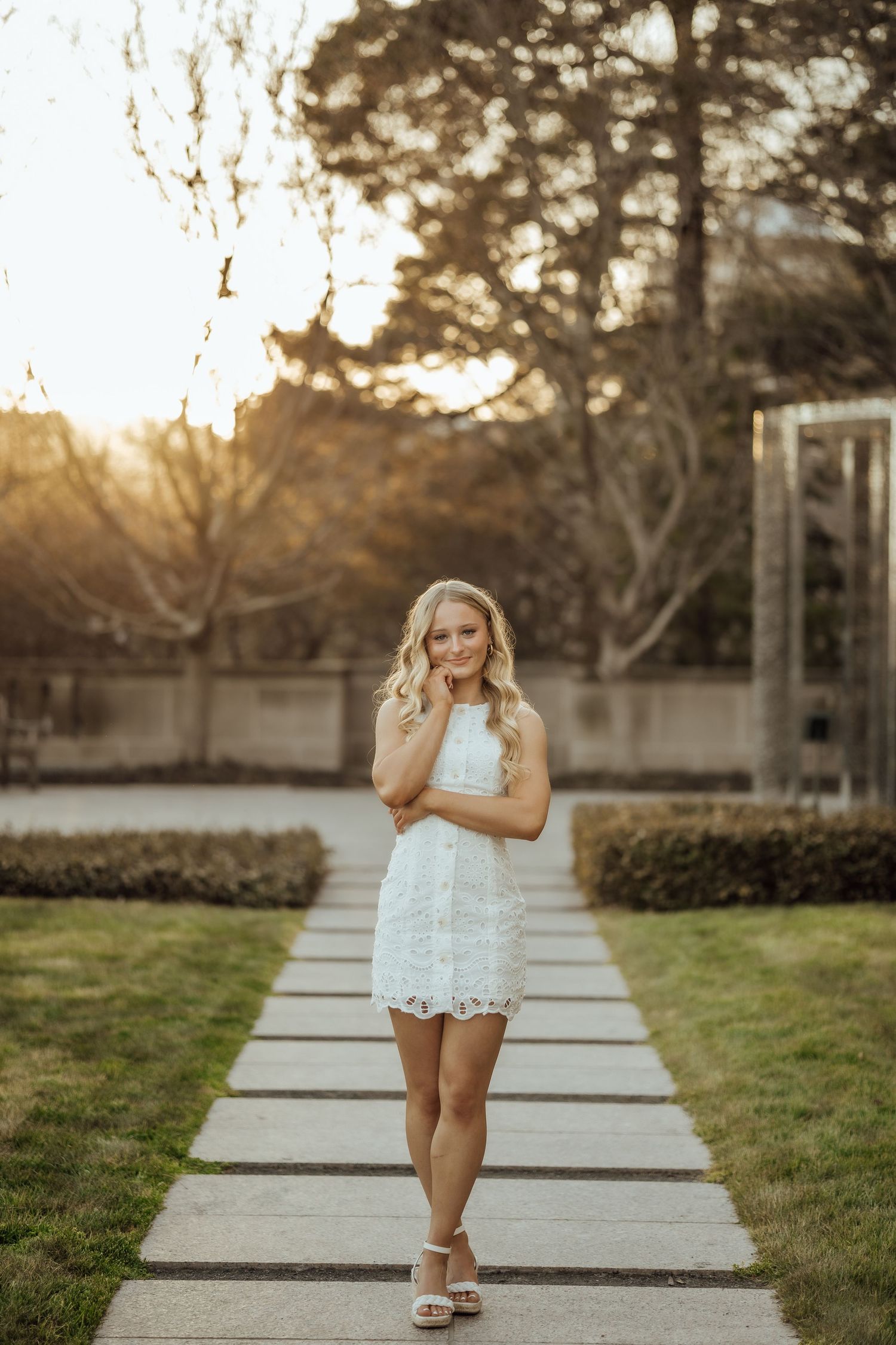 Smiling blonde woman in white lace mini dress posing on garden pathway in warm sunset light.