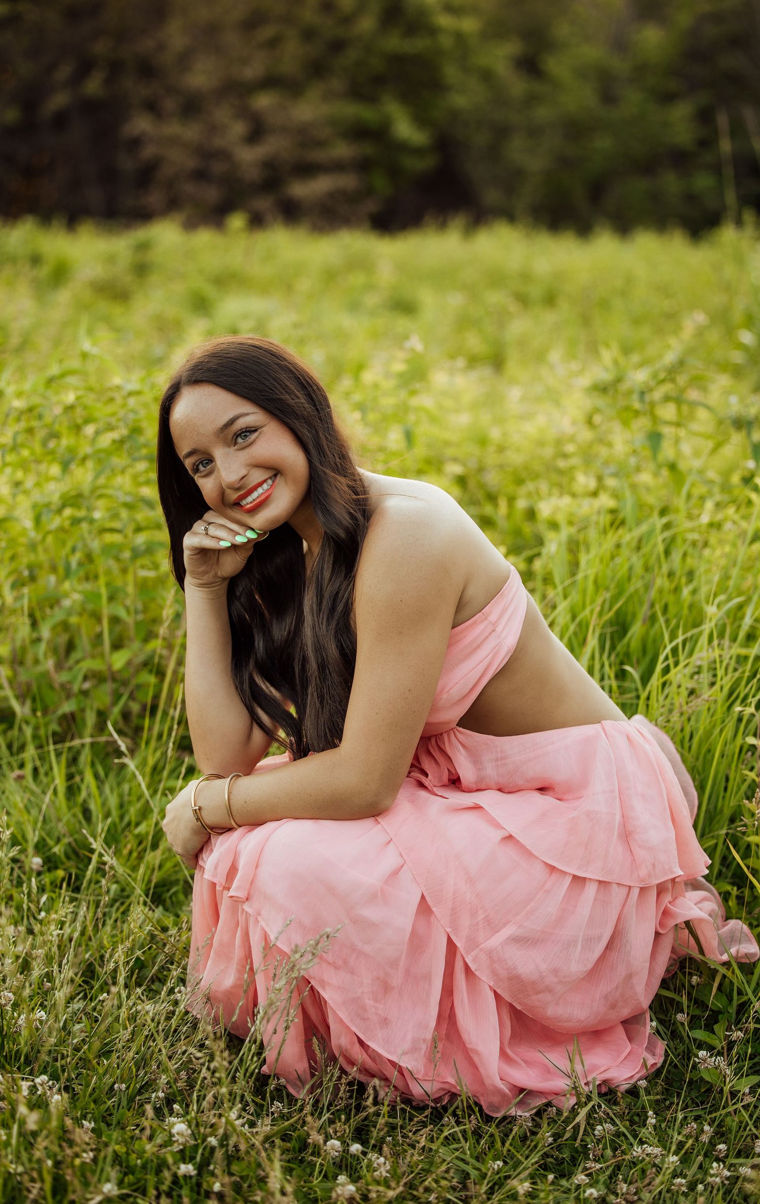 Smiling young woman in pink ruffled dress sitting in a green wildflower meadow.