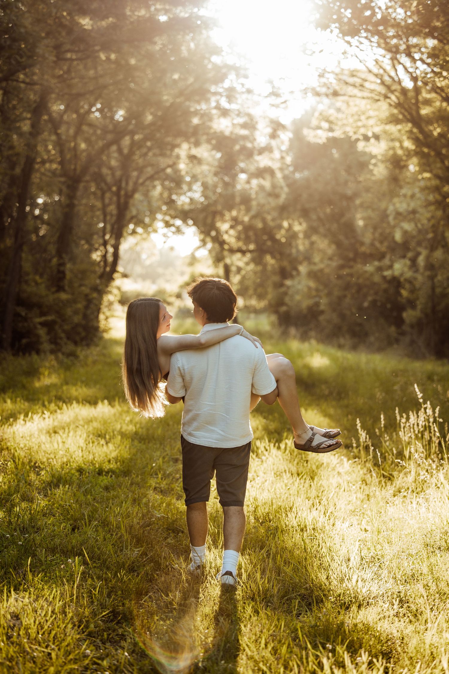 A boy carries a girl piggyback through a sunlit forest path surrounded by lush green trees and golden light.