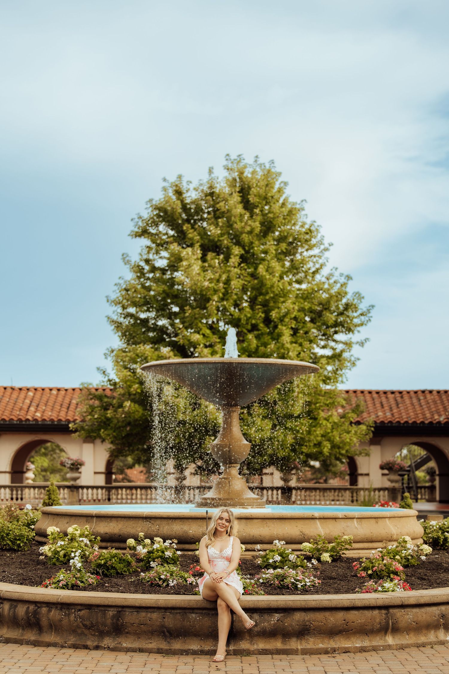 A woman in a white dress sits gracefully by an ornate stone fountain surrounded by flowers and lush greenery.