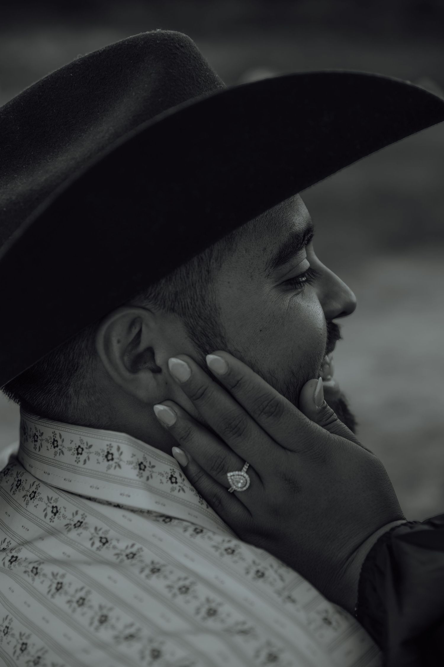 Black and white close-up profile of man in cowboy hat with woman's hand touching his face showing engagement ring.