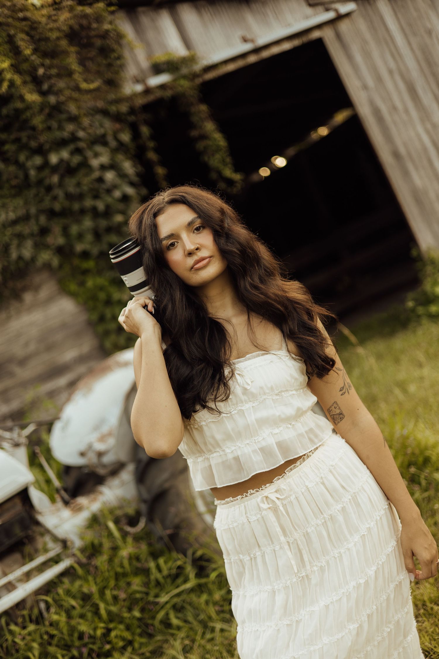 Young woman in white ruffled crop top and skirt holds camera outdoors near rustic barn with ivy.