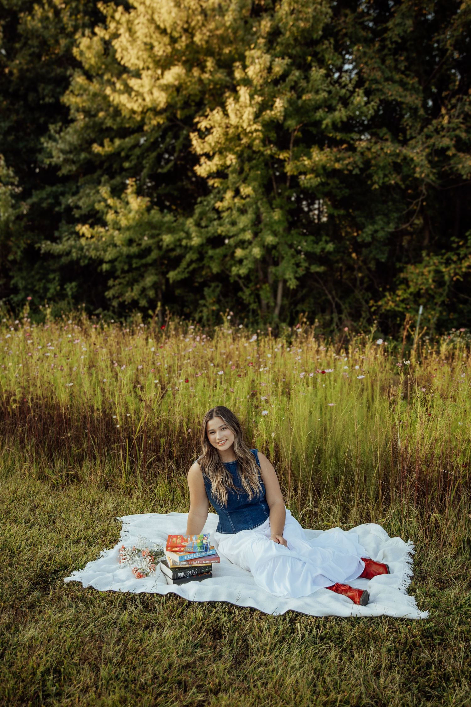 Smiling brunette woman sitting on white blanket in meadow with stack of colorful books at sunset.