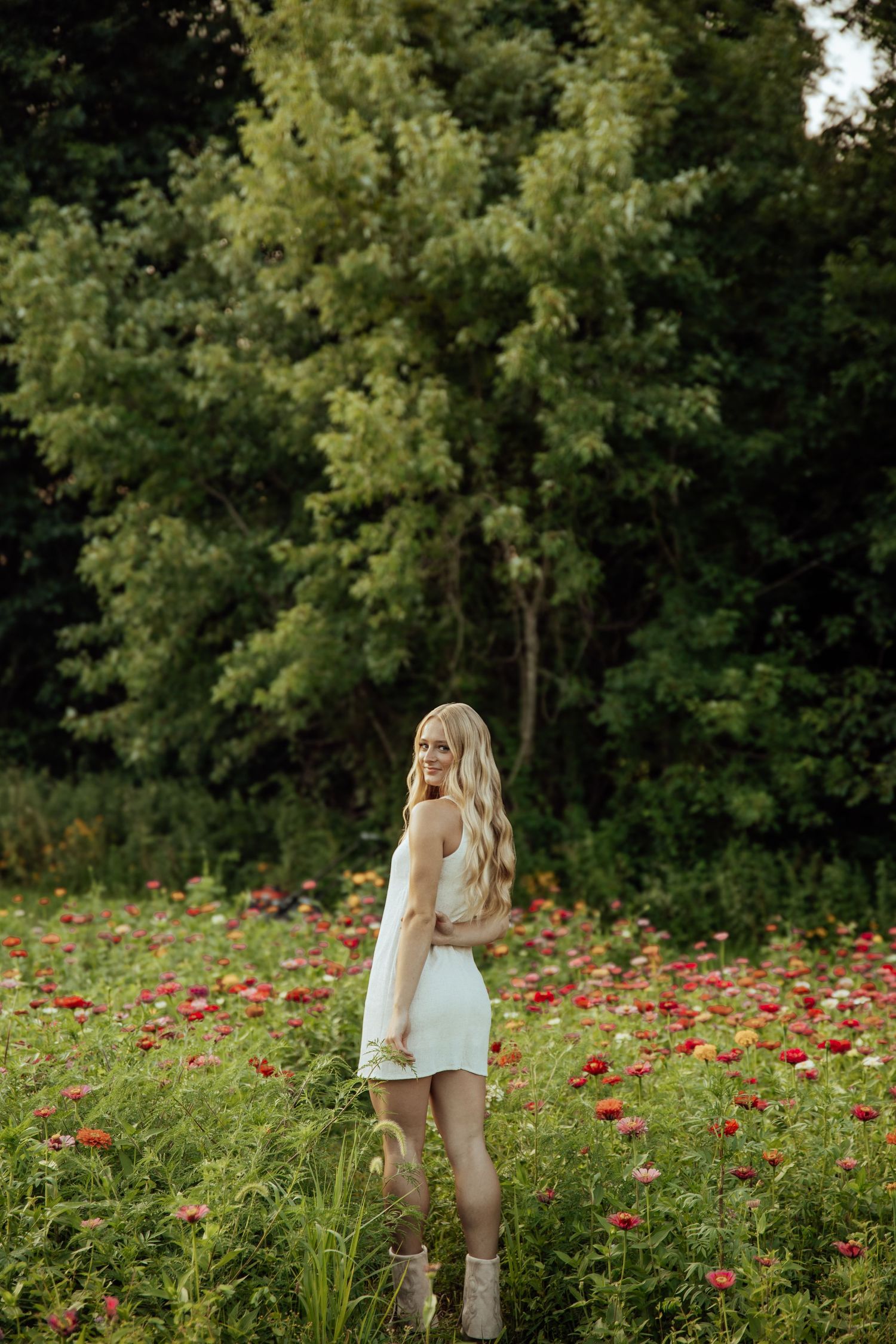 Blonde woman with long wavy hair in white mini dress standing in red flower field near trees.