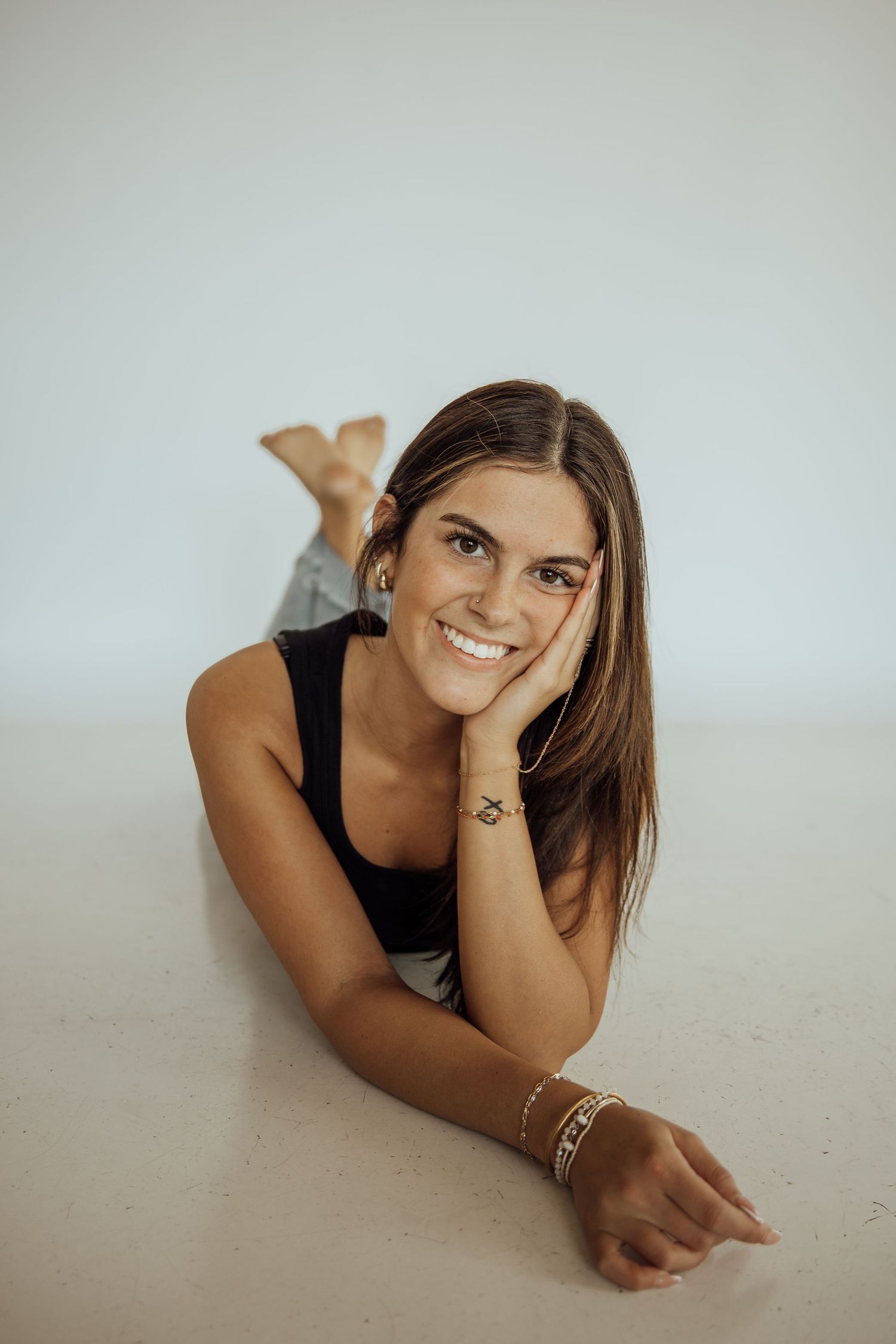 Smiling young woman in black tank top lying on white floor with hand resting on cheek.