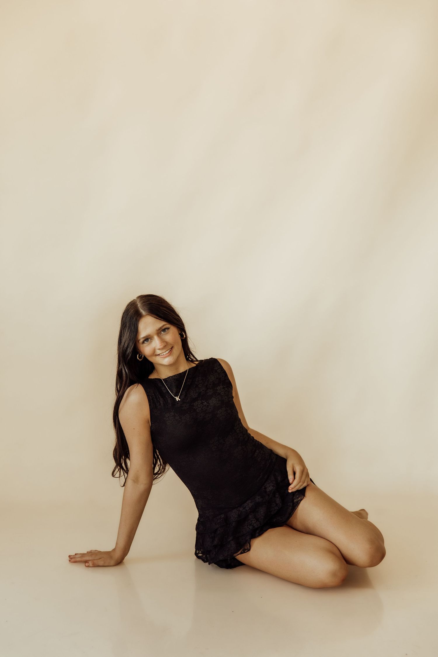 Young woman in black sleeveless mini dress sitting on floor, smiling, against beige background.