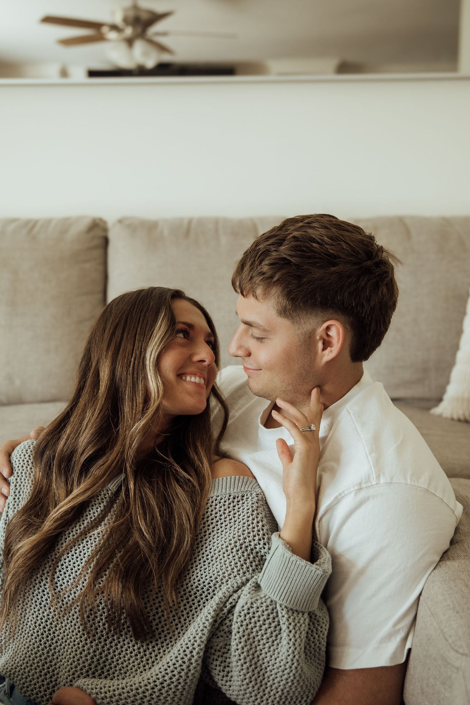 Young couple sitting against a couch indoors, smiling at each other in a cozy home setting.