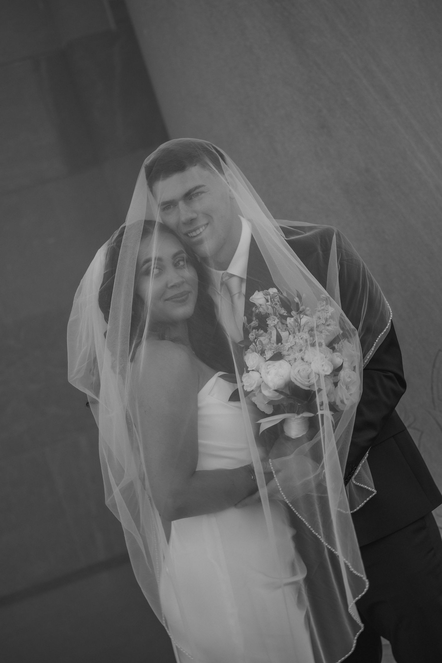 Black and white portrait of smiling newlyweds sharing a romantic moment under bride's wedding veil.