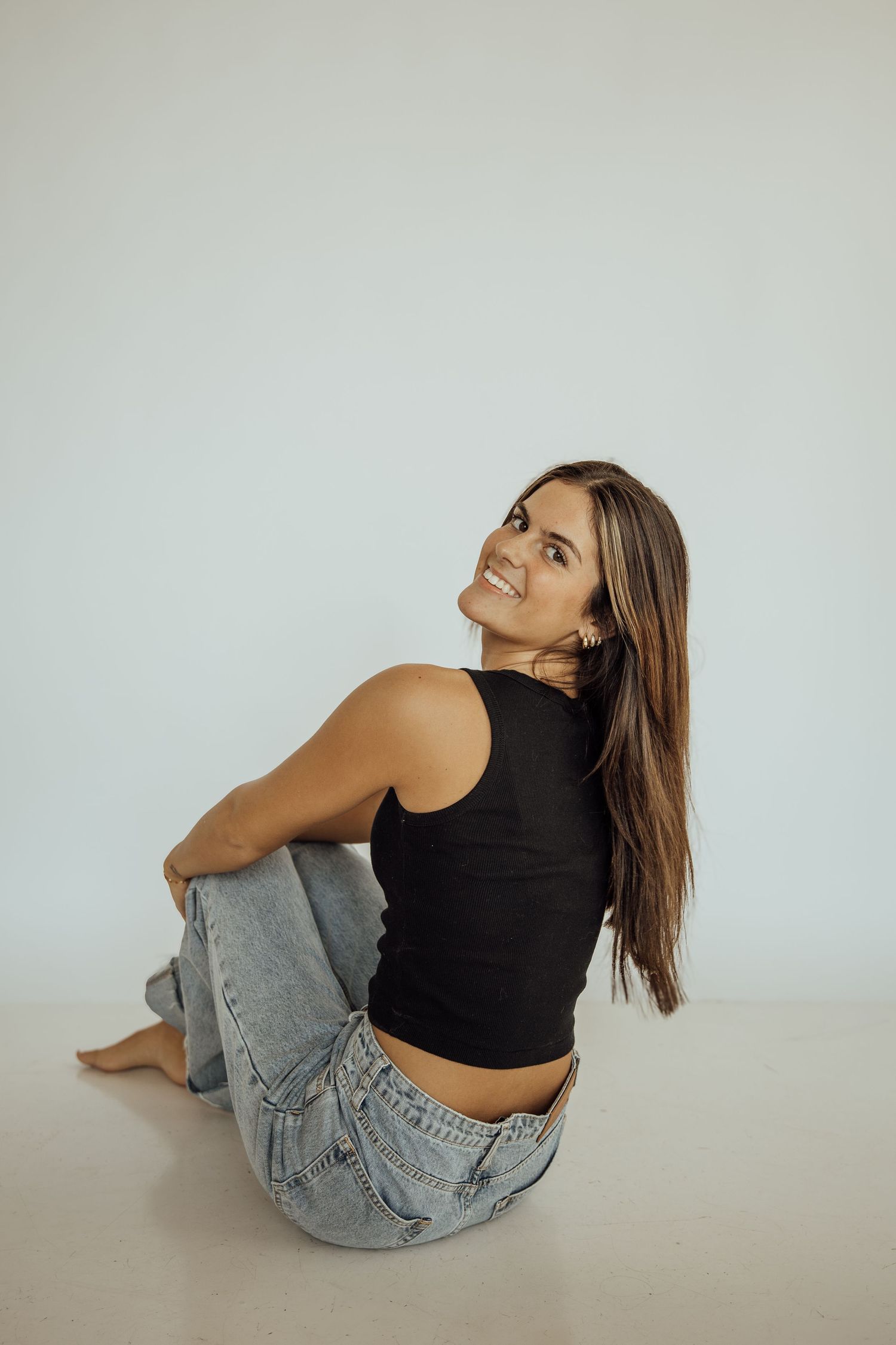 Young woman in black tank top and jeans sitting barefoot on white floor looking upward.