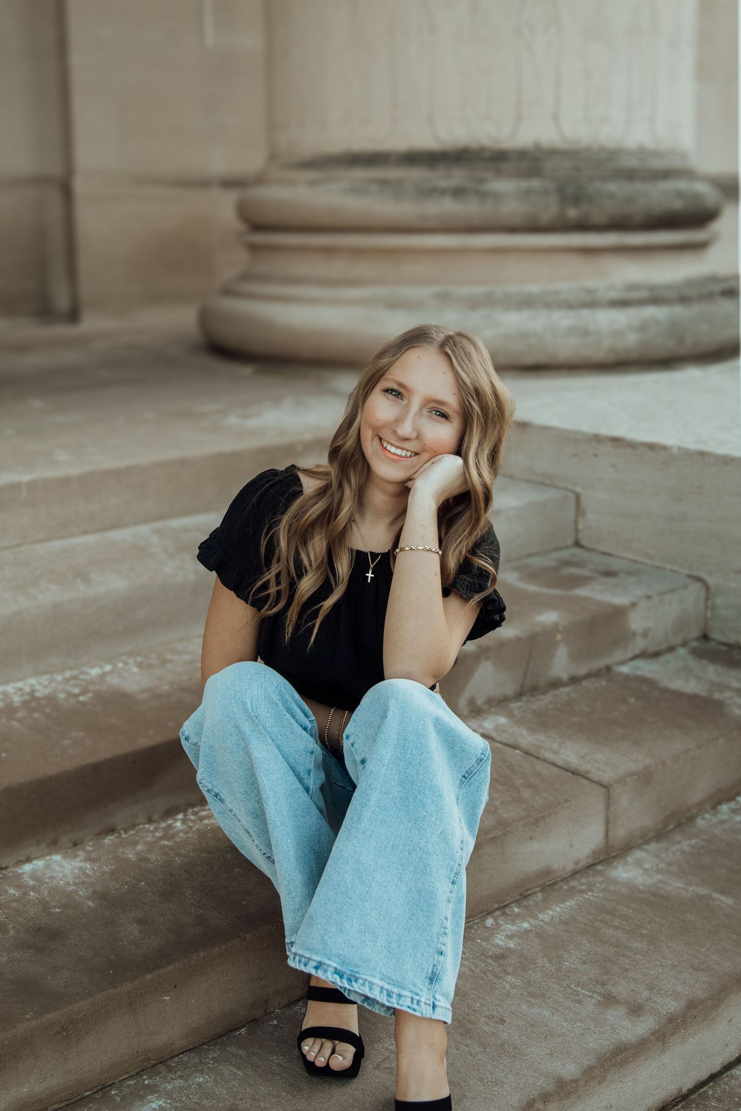 Smiling young woman in black top and light wash jeans sitting on stone steps by a column.