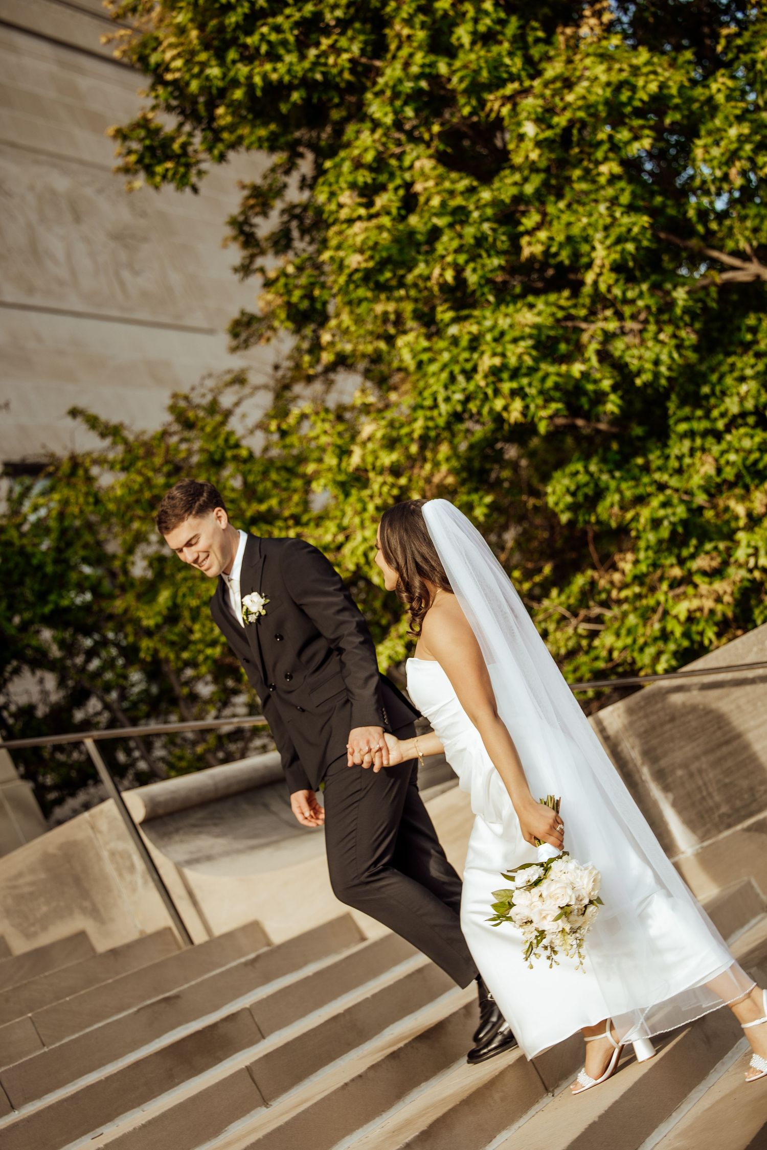 Newlywed couple holding hands while walking up outdoor stone steps surrounded by green trees.