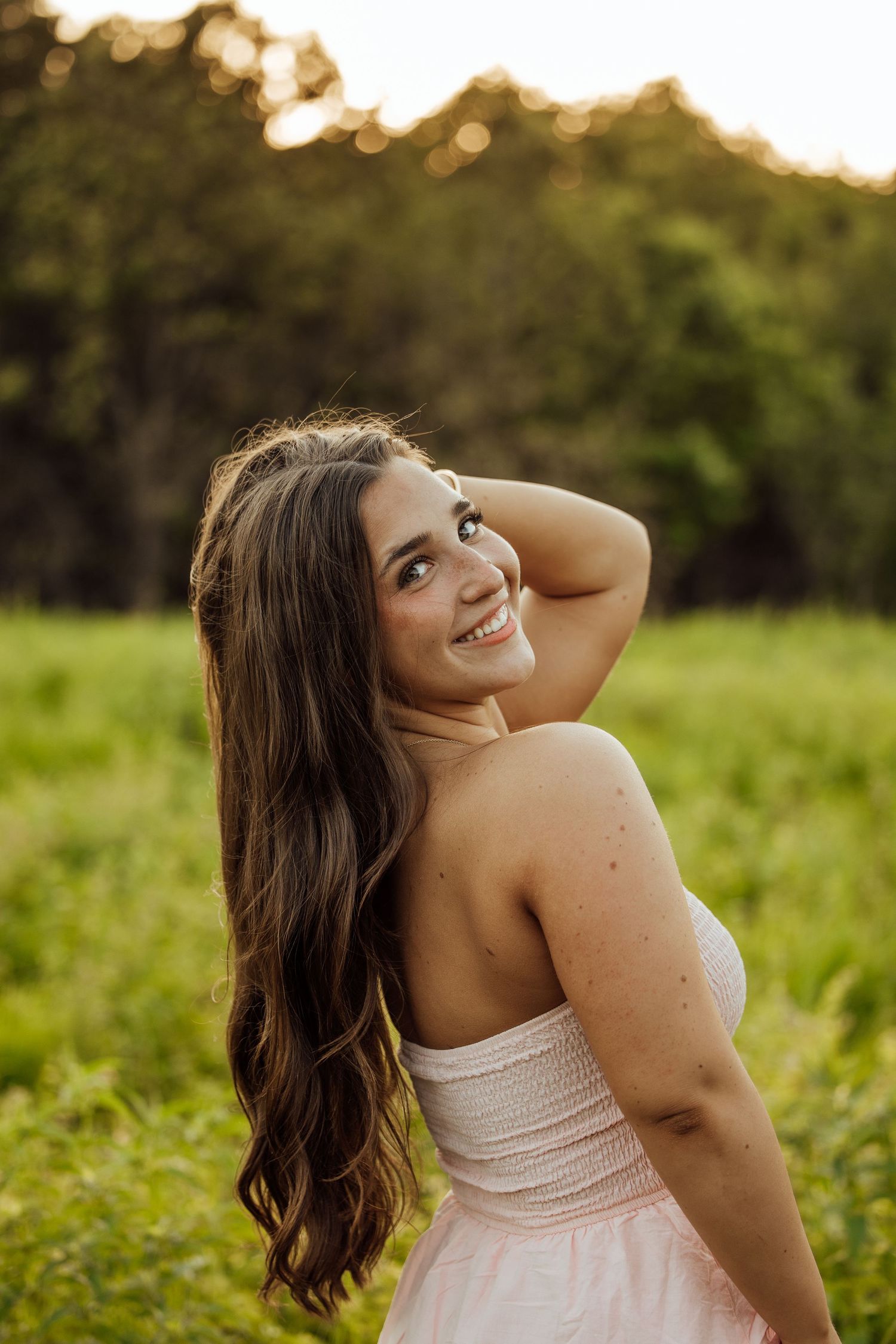 Beautiful brunette woman in pink dress smiling over shoulder in golden hour sunlit green field.