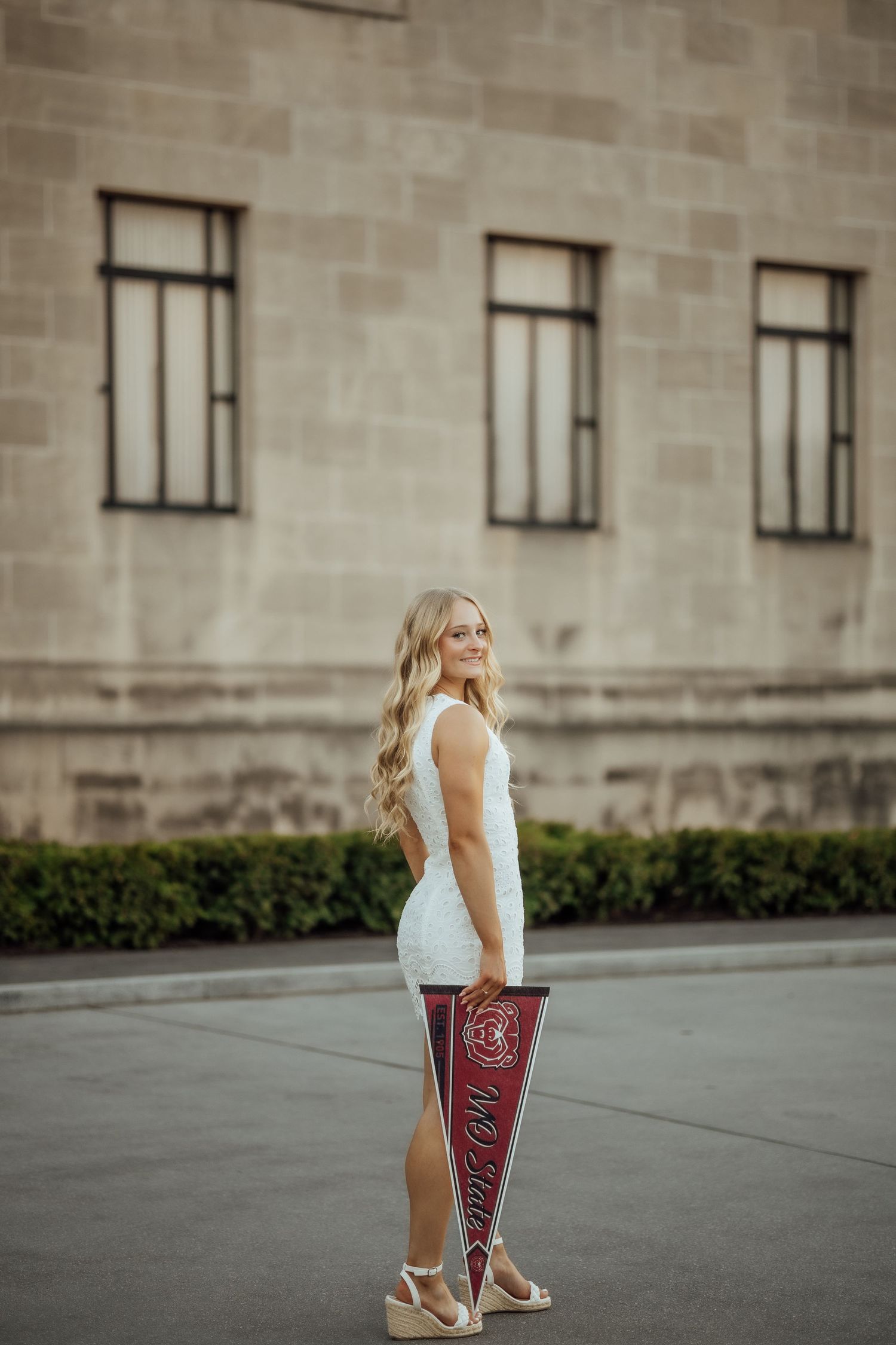 Blonde woman in white lace dress holds MO State pennant beside stone building.