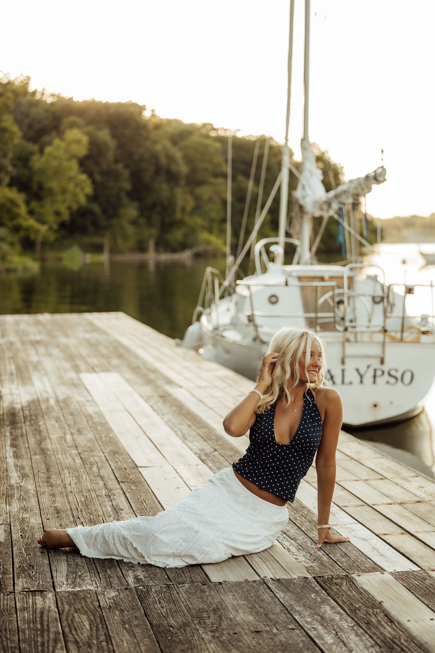 Blonde woman in polka dot top and white skirt sitting on wooden dock near sailboat named Calypso at sunset.